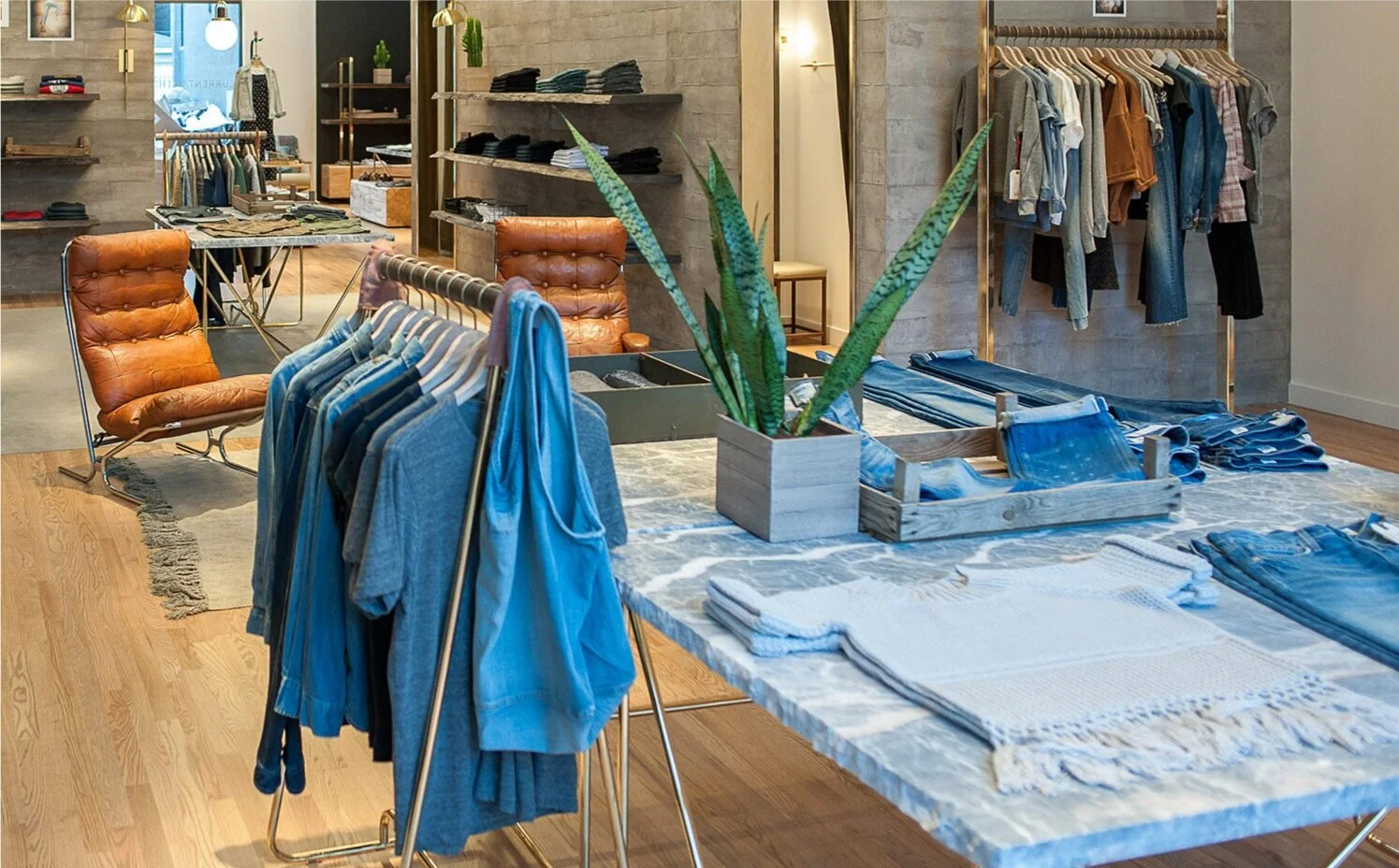 Denim clothing and a plant on a marble table display inside a retail store with camel colored chairs in the background. To the right are women's denim clothing on a gold colored rack.