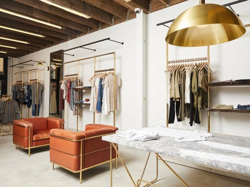 Retail clothing space with two brown distressed leather chairs. A brass lamp hangs from the exposed ceiling beams. Brass clothing racks with clothes are on the back wall.