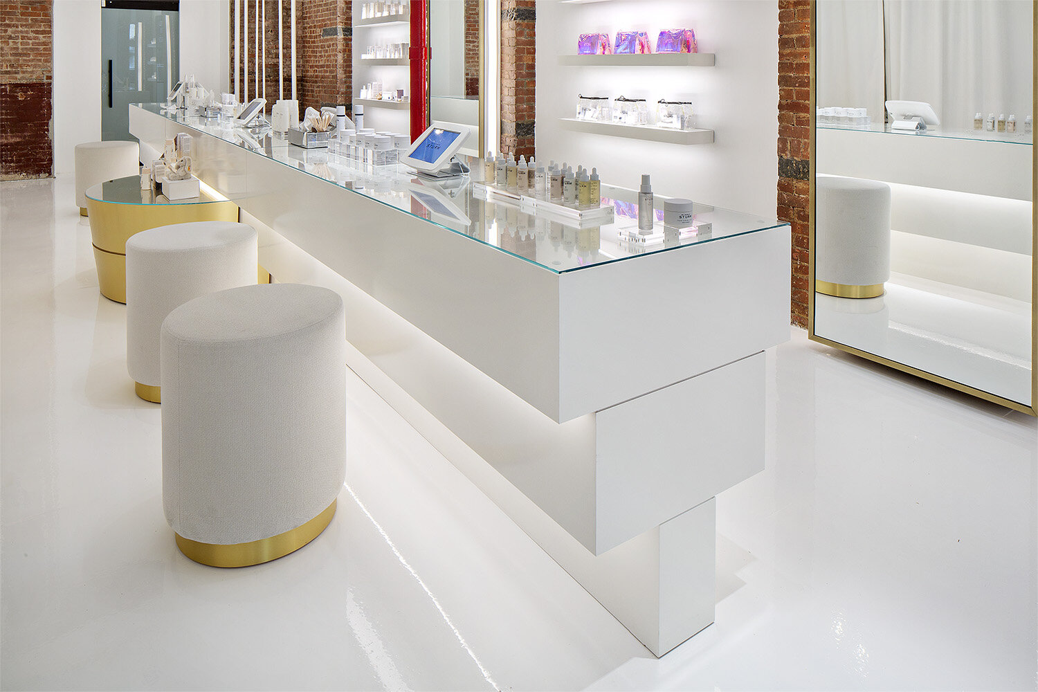 A white skincare counter with a glass tabletop with product displayed. Round white stools with gold accents are tucked under the counter.