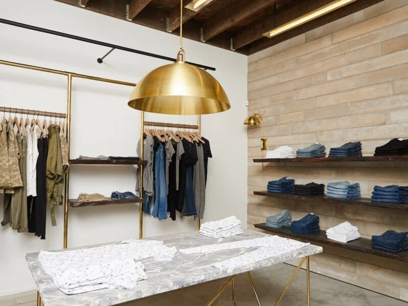 Shiny brass lighting fixture hangs from exposed ceiling beams in a retail clothing store. A marble table features white clothing, and the back wall shows clothing being shown on racks and floating shelves.