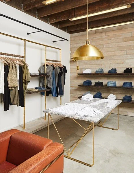 Retail clothing space with a brown distressed leather chair. A brass lamp hangs from the exposed ceiling beams. Brass clothing racks with clothes are on the back wall.