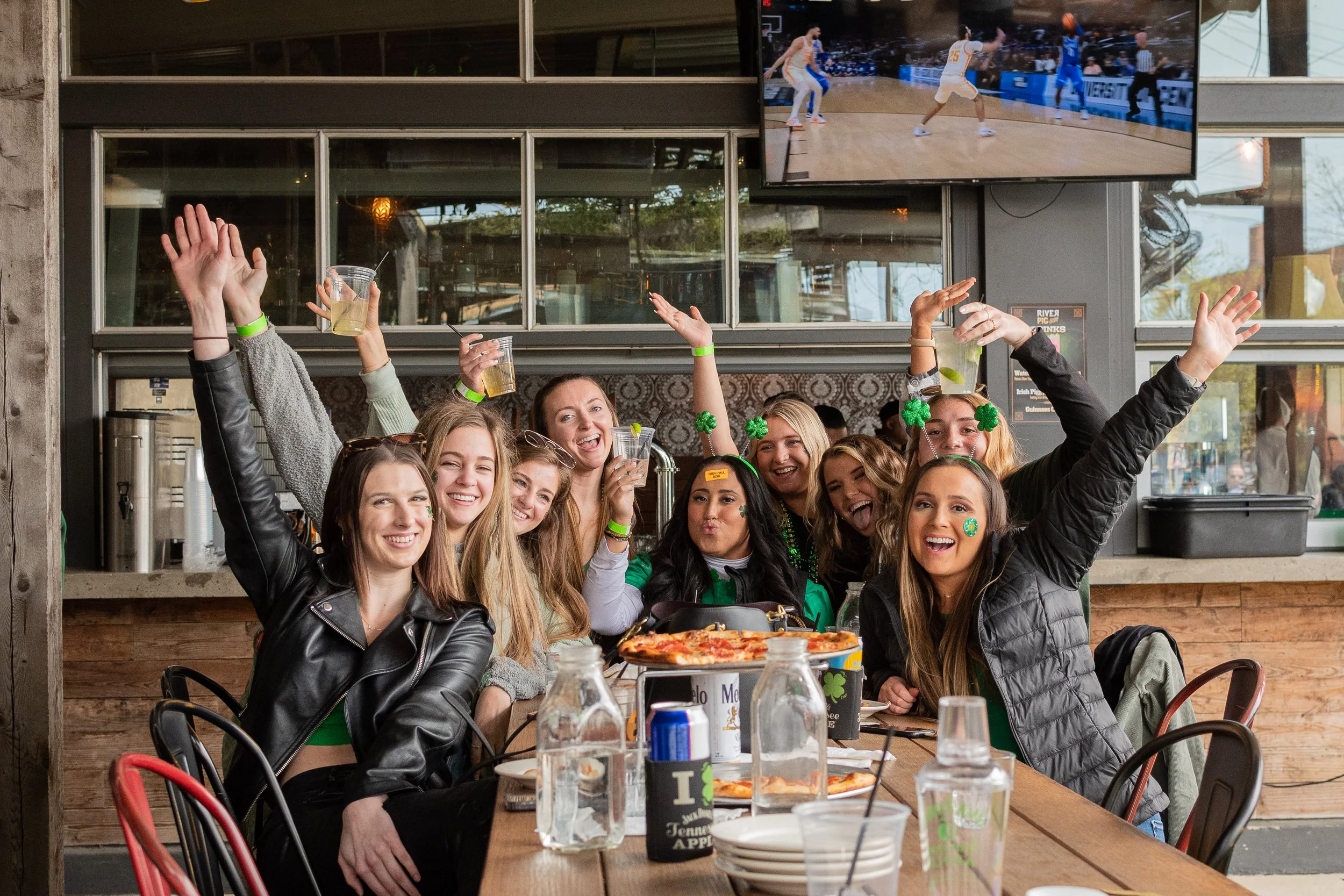 Group of young women celebrating at a restaurant, with some raising drinks and smiling