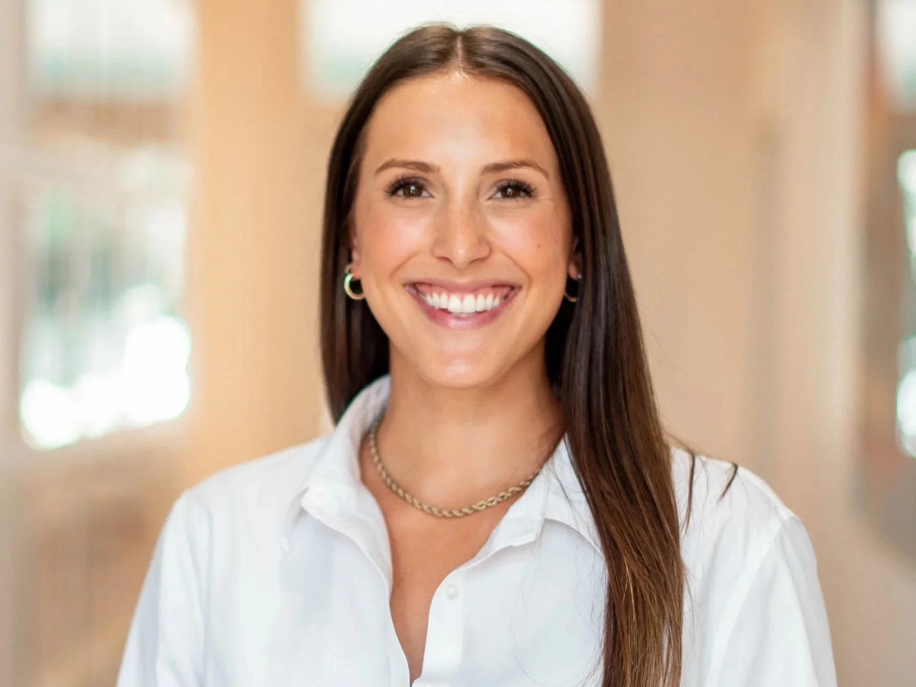 Natalie Draeger smiling and wearing a white button-up shirt, standing indoors in front of windows.