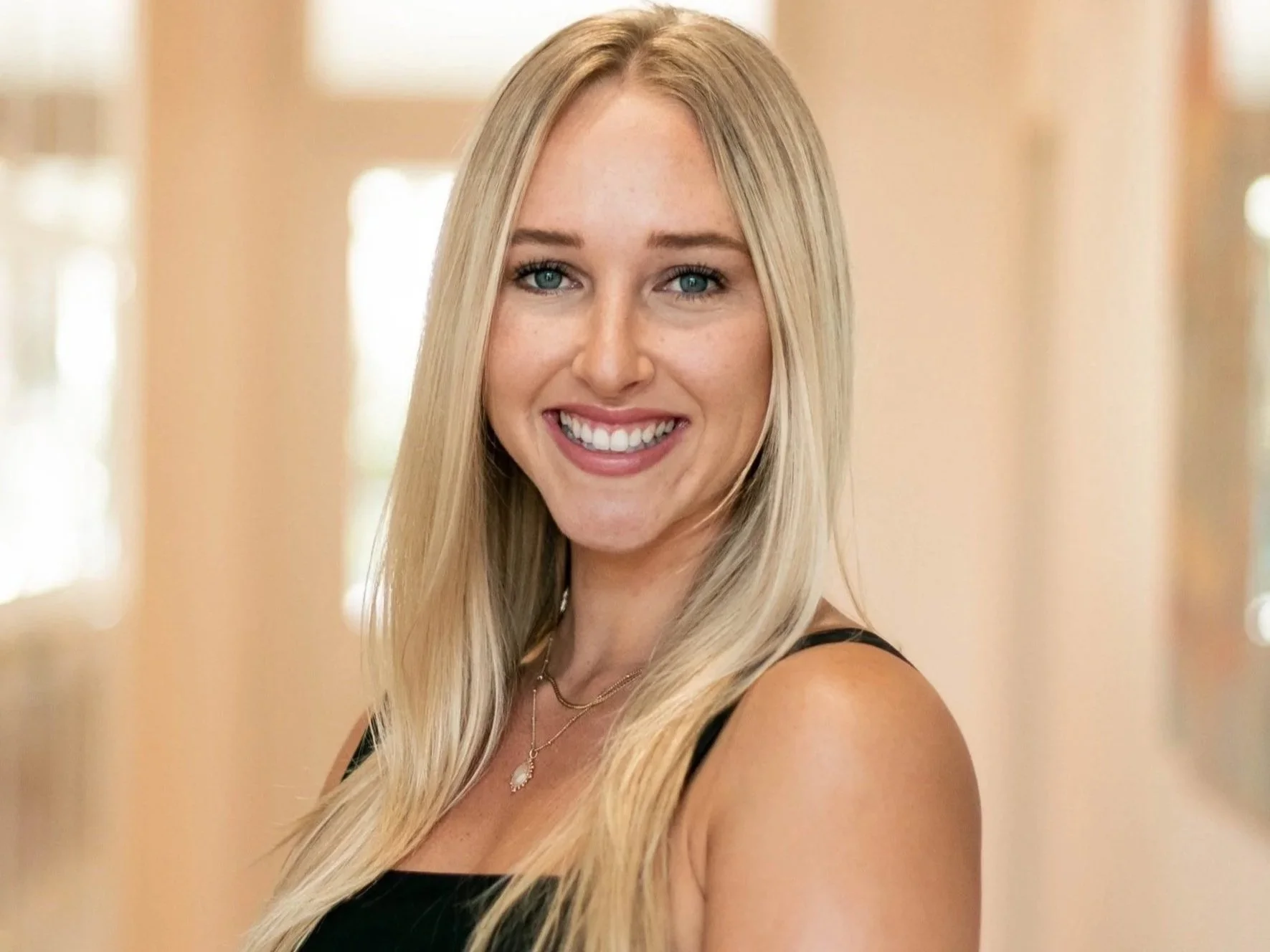 Anna McSwiggen smiling and wearing a black sleeveless dress and jewelry, standing indoors.