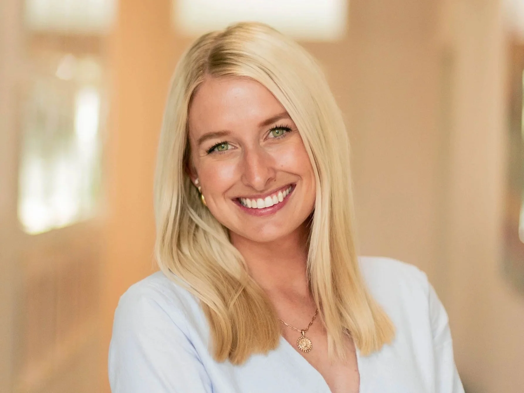 Jenna Waters smiling and wearing a light blue blouse, gold jewelry, and standing in a well-lit hallway.