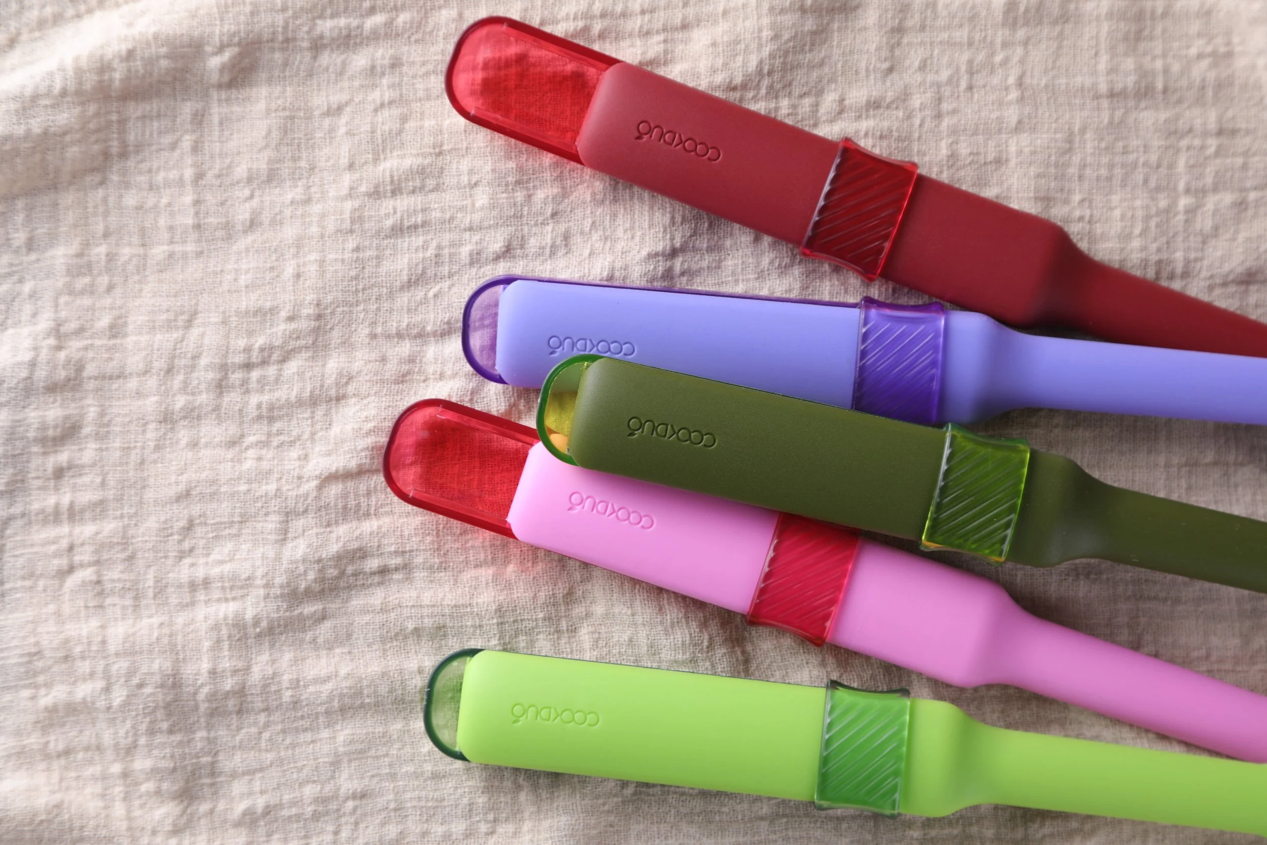 Five Cookduo Mix and Measure silicone spoons fanned on linen in red, purple, olive, pink, and lime colorways with concealed measuring spoon caps
