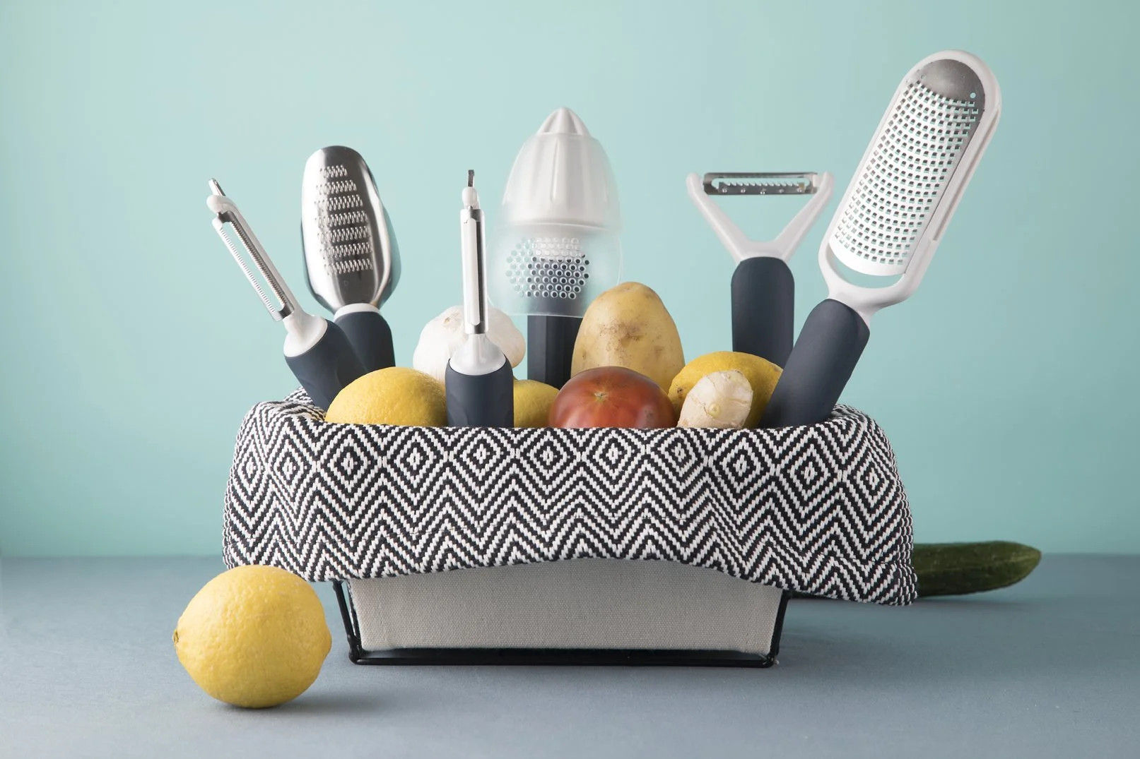 Cookduo Food Prep Line collection displayed in a chevron cloth basket with fresh lemons, ginger, potato, and tomato against a mint-blue wall