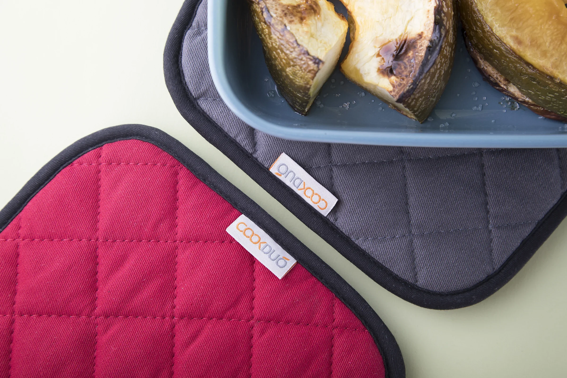 Cookduo Grab and Grip pot holders in red and gray seen top-down with a baking pan of roasted squash on a sage kitchen surface