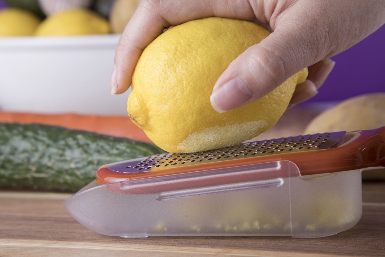 Hand zesting a lemon with the Cookduo Food Prep zester, showing the integrated collection and pour container that catches citrus zest cleanly