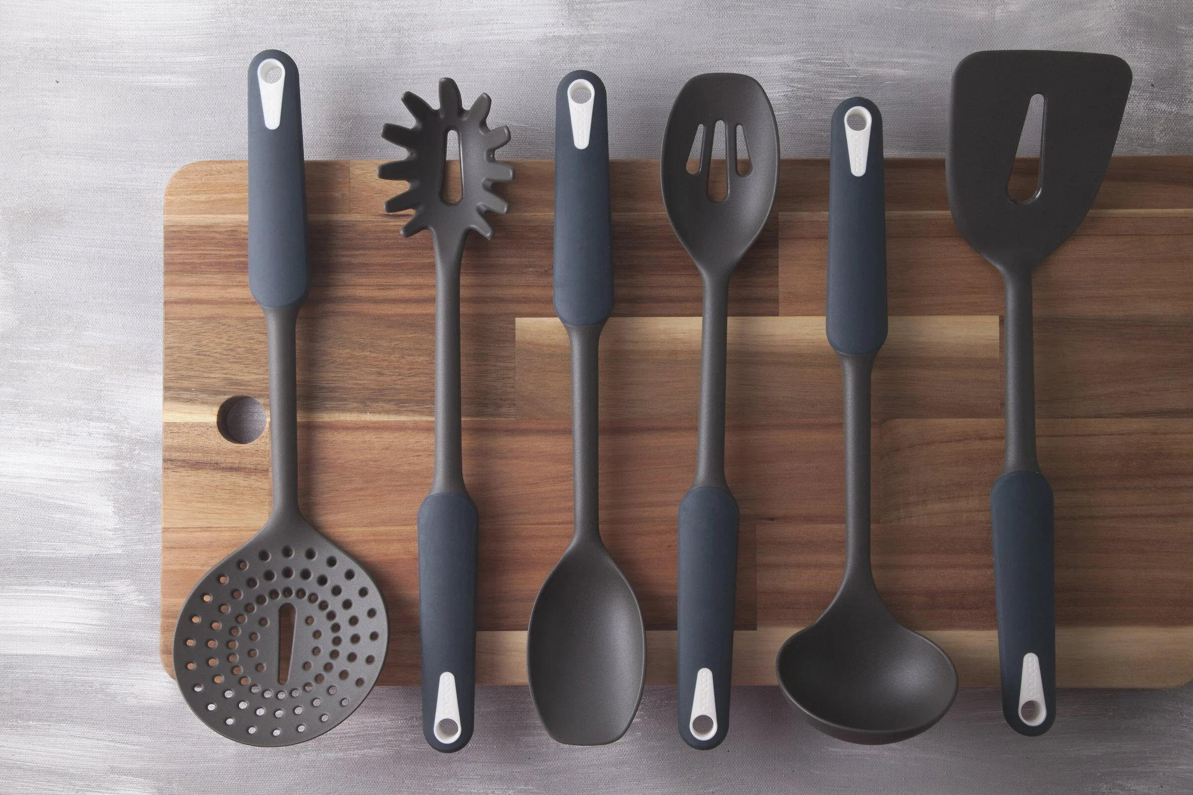Cookduo Steelcore Nylon Tools arranged on a wooden cutting board: skimmer, pasta server, solid spoon, slotted spoon, ladle, and slotted turner