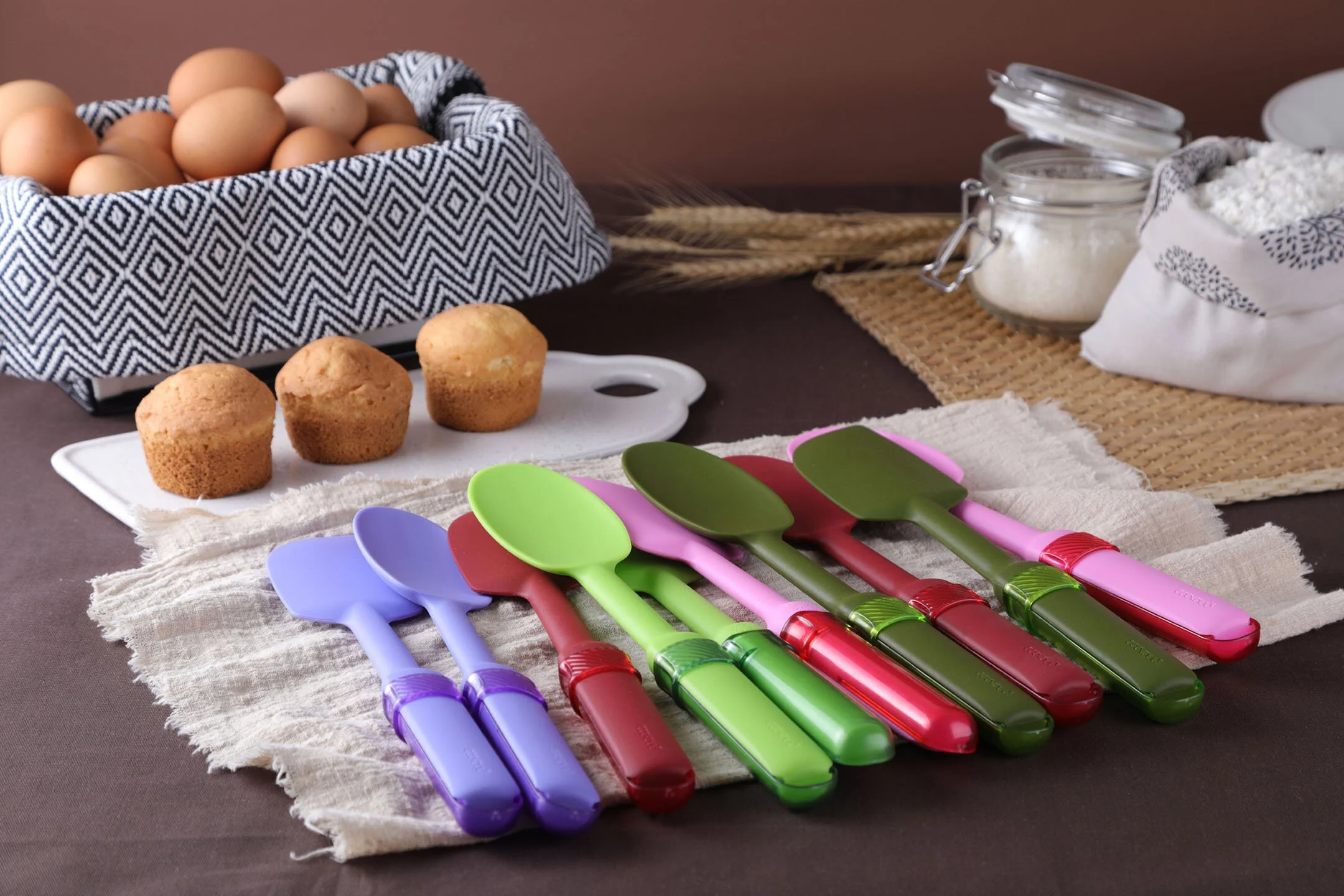 Cookduo Mix and Measure silicone tools arranged in a baking scene with muffins, eggs, sugar, and flour, showing the two-in-one spoon and measure design