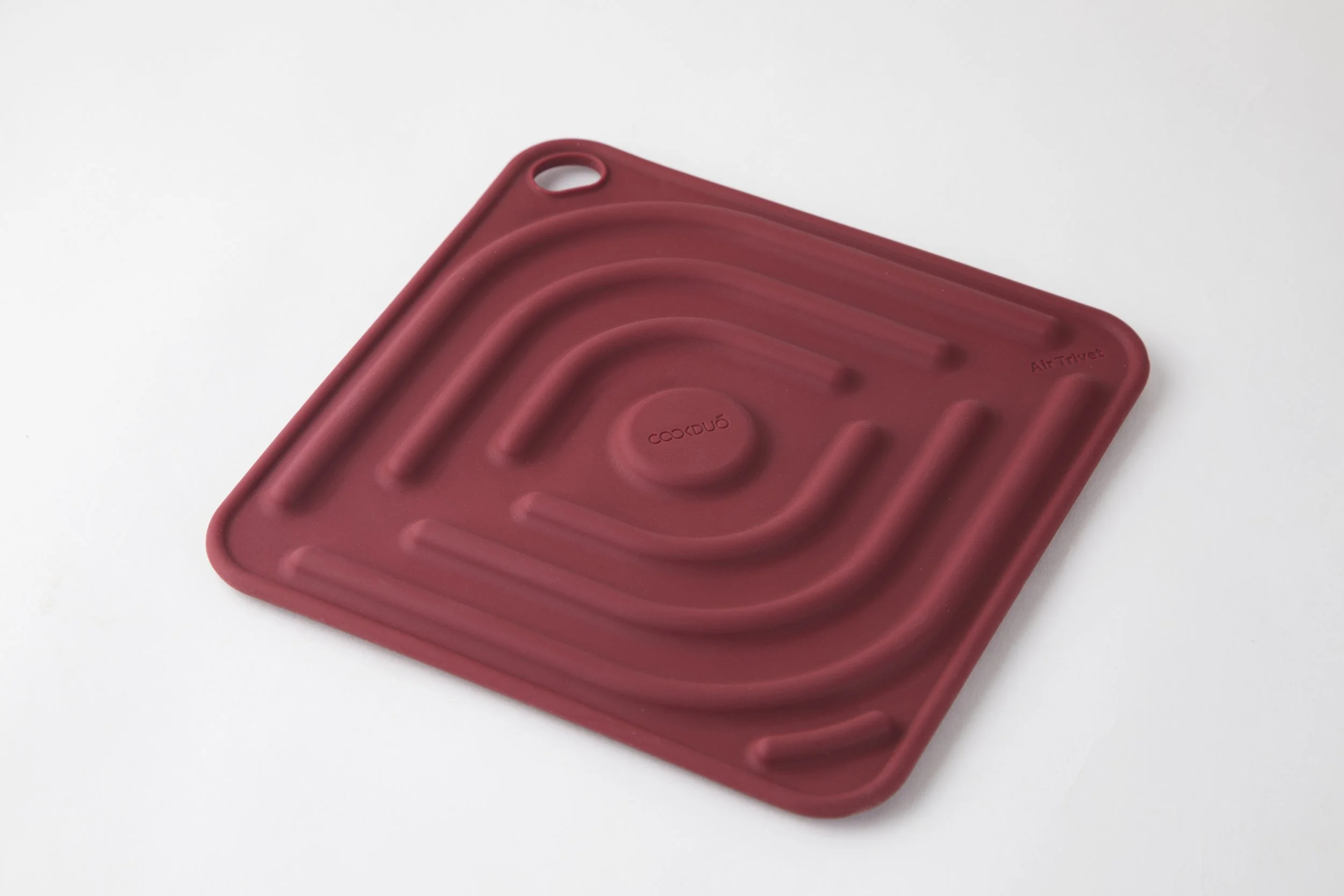 Single Cookduo Air Trivet in burgundy at a 3/4 angle on white, showing the silicone heat-protection trivet with debossed logo