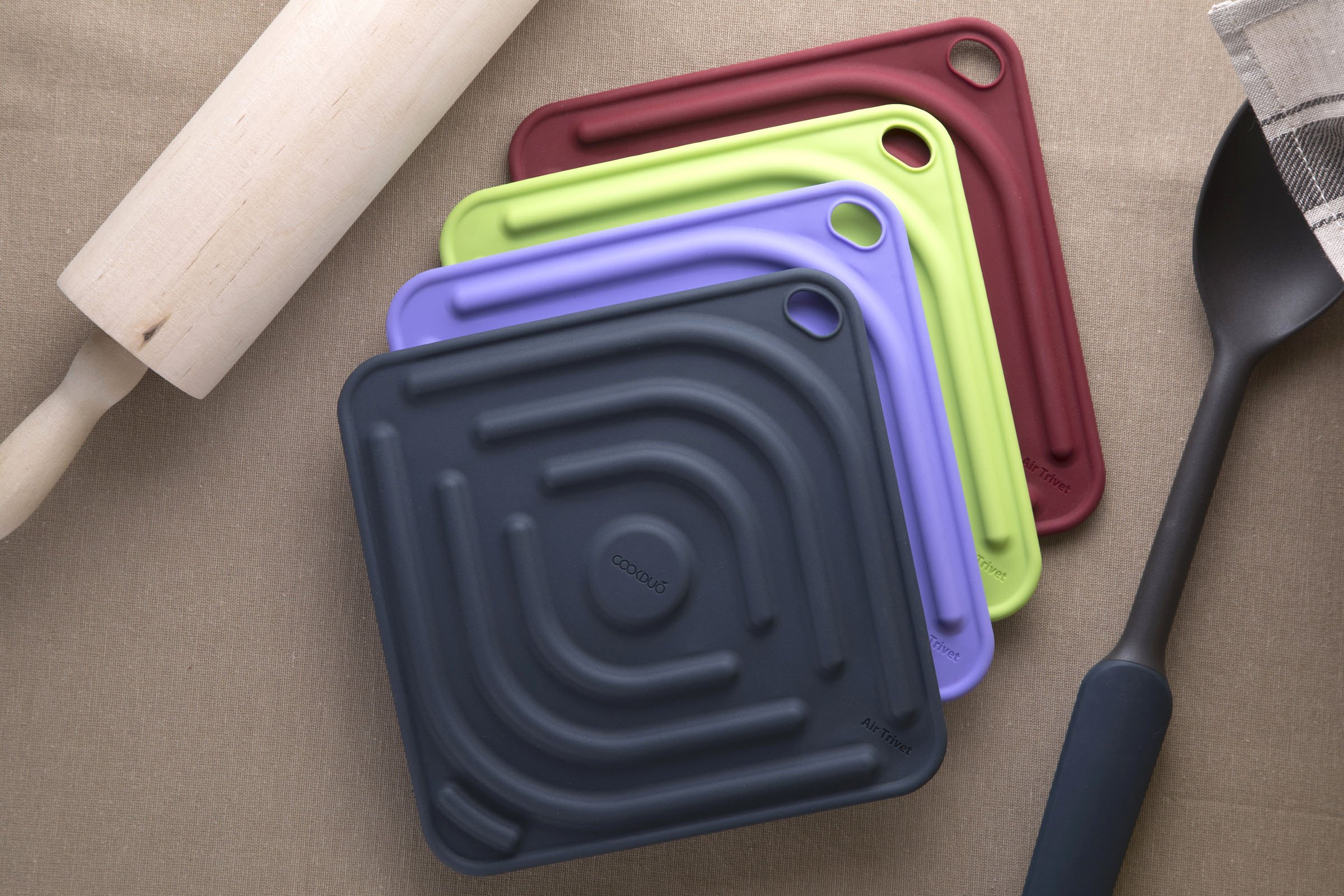 Cookduo Air Trivet silicone trivets in navy, lime, purple, and burgundy fanned on a tabletop with a rolling pin and spatula