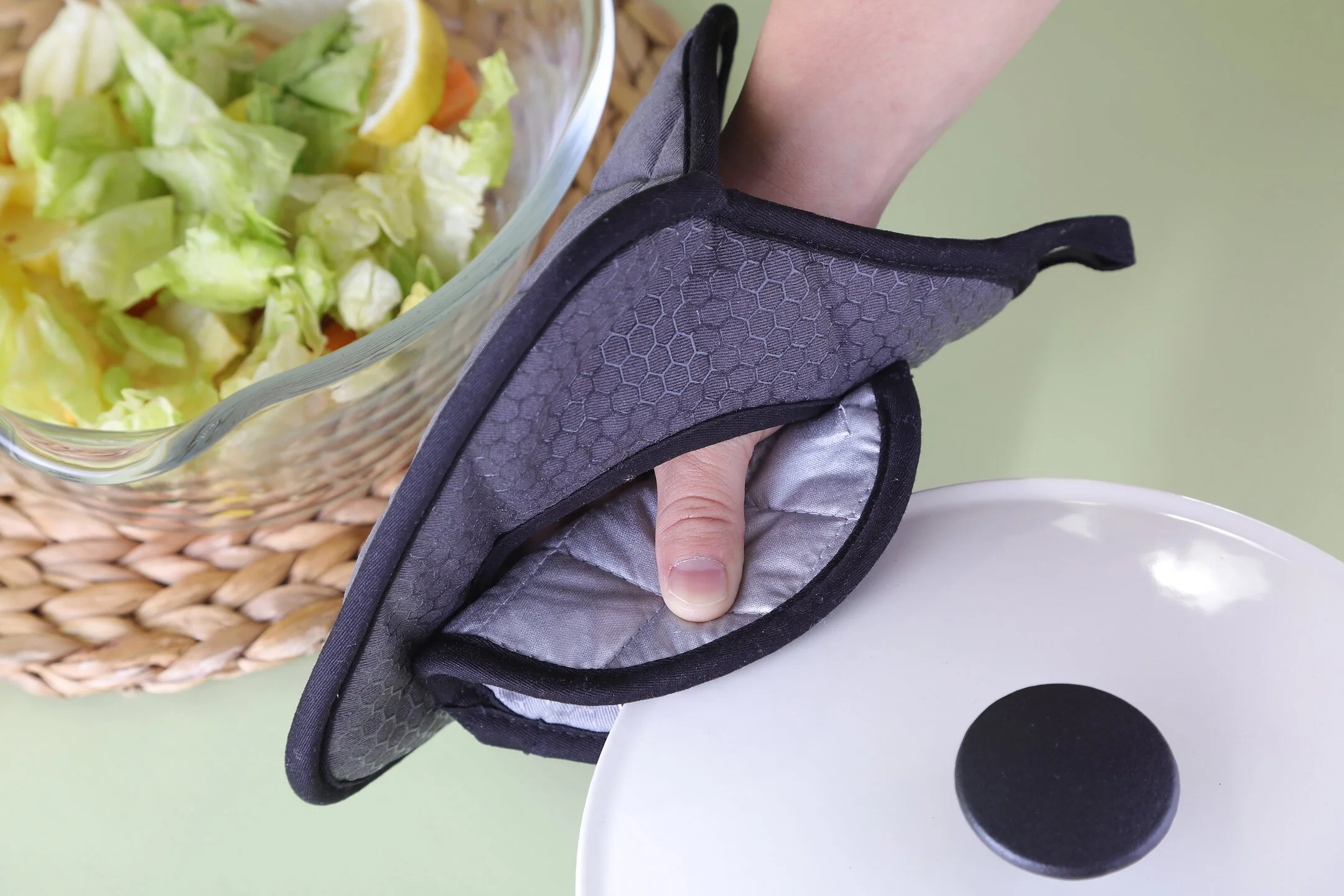 Hand inserted in the mitt pocket of a Cookduo Grab and Grip pot holder, lifting a hot pot lid next to a fresh salad bowl