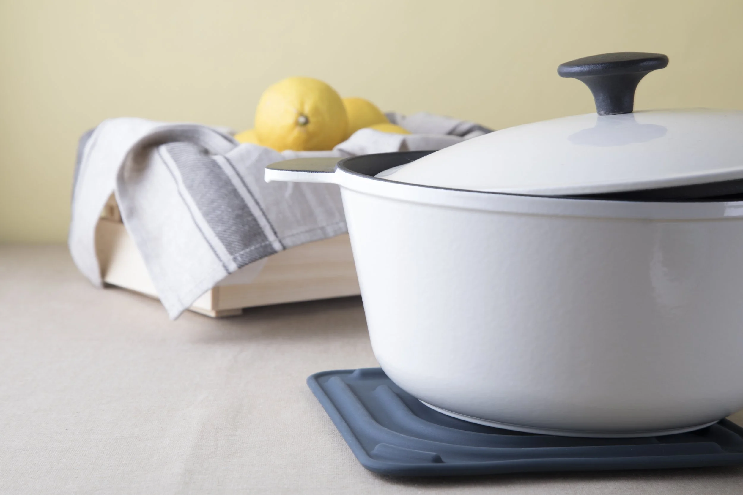 White enamel Dutch oven resting on a navy Cookduo Air Trivet with fresh lemons beside it, showing the trivet protecting the kitchen table from hot cookware