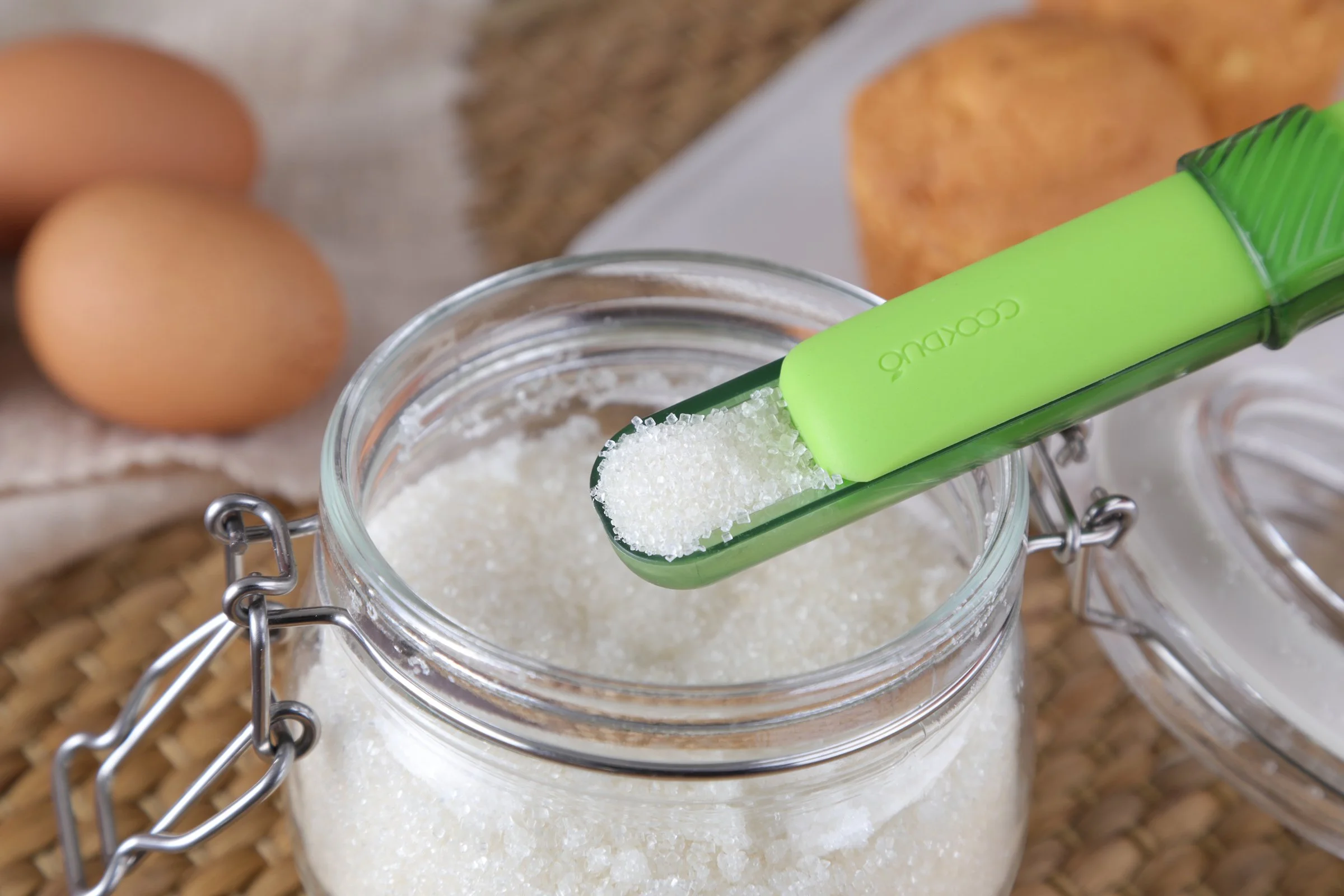 Green Cookduo Mix and Measure silicone spoon scooping sugar from a glass jar during baking, with brown eggs and muffins in the background