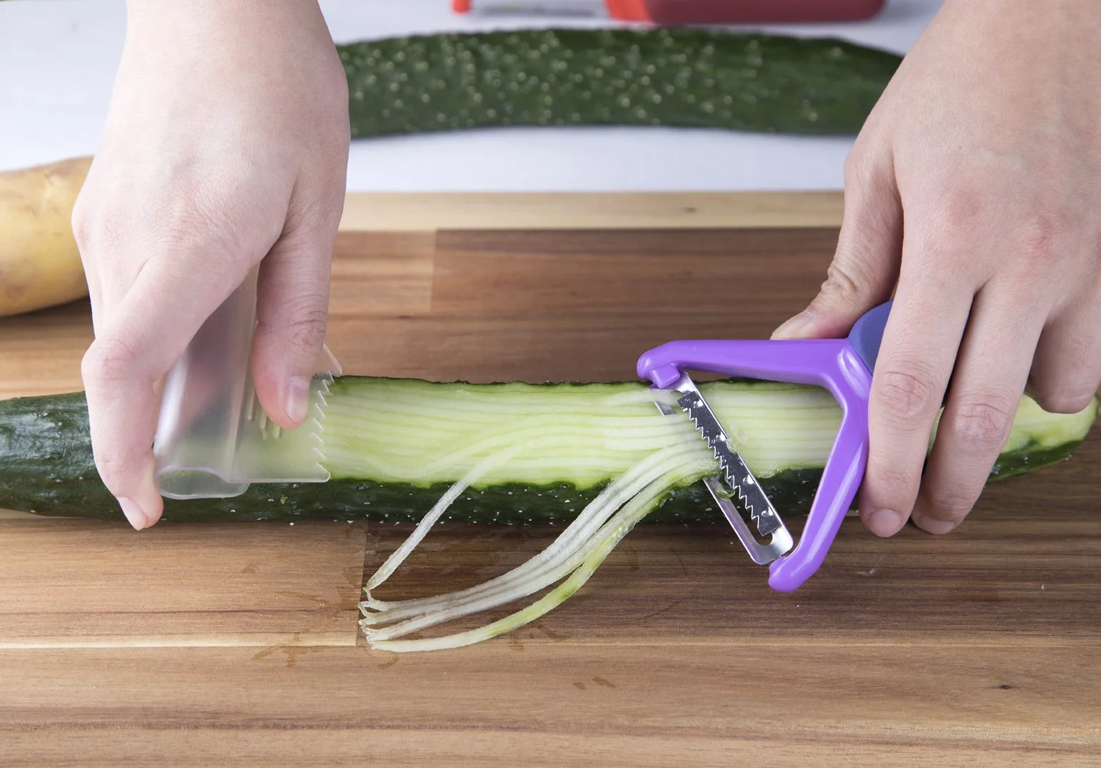 Hands using the purple Cookduo Food Prep julienne peeler with integrated safety guard to make long julienne strips from a cucumber