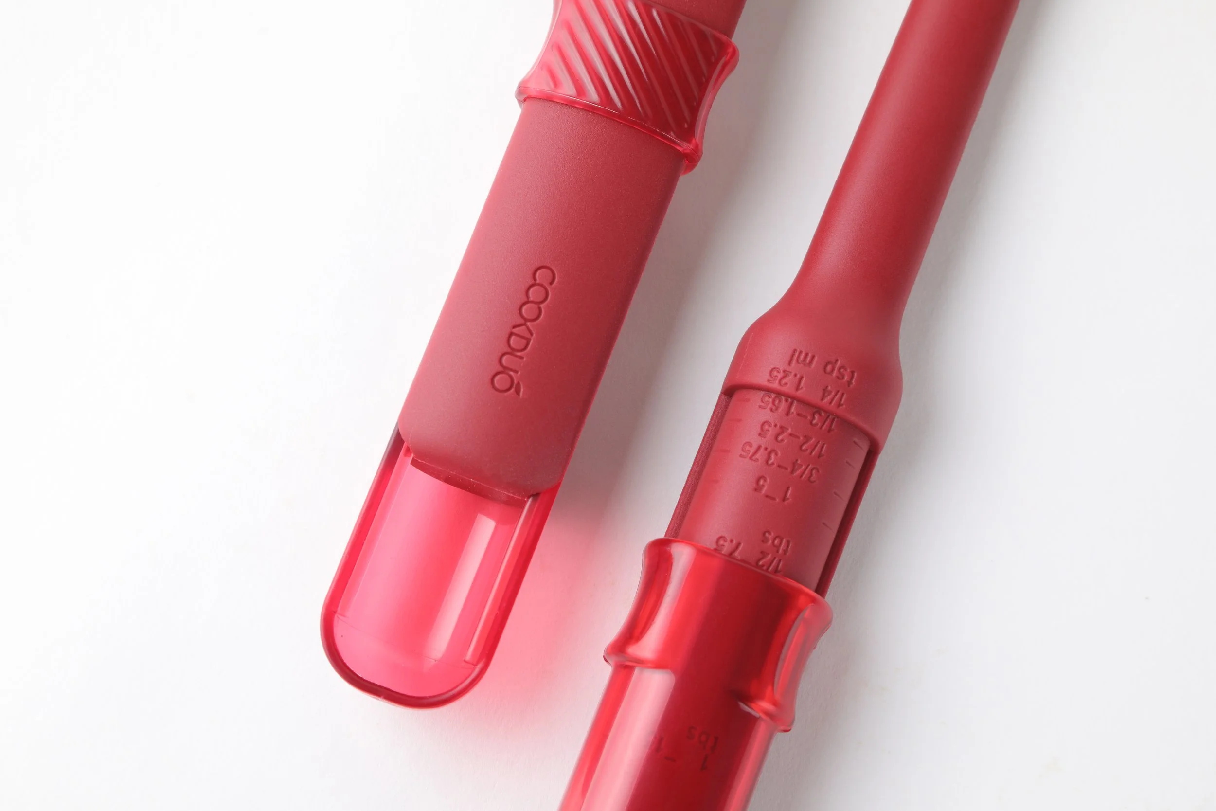 Close-up of the Cookduo Mix and Measure silicone spoon showing the built-in sliding measuring spoon and volume markings debossed in the handle in tbs, tsp, and ml