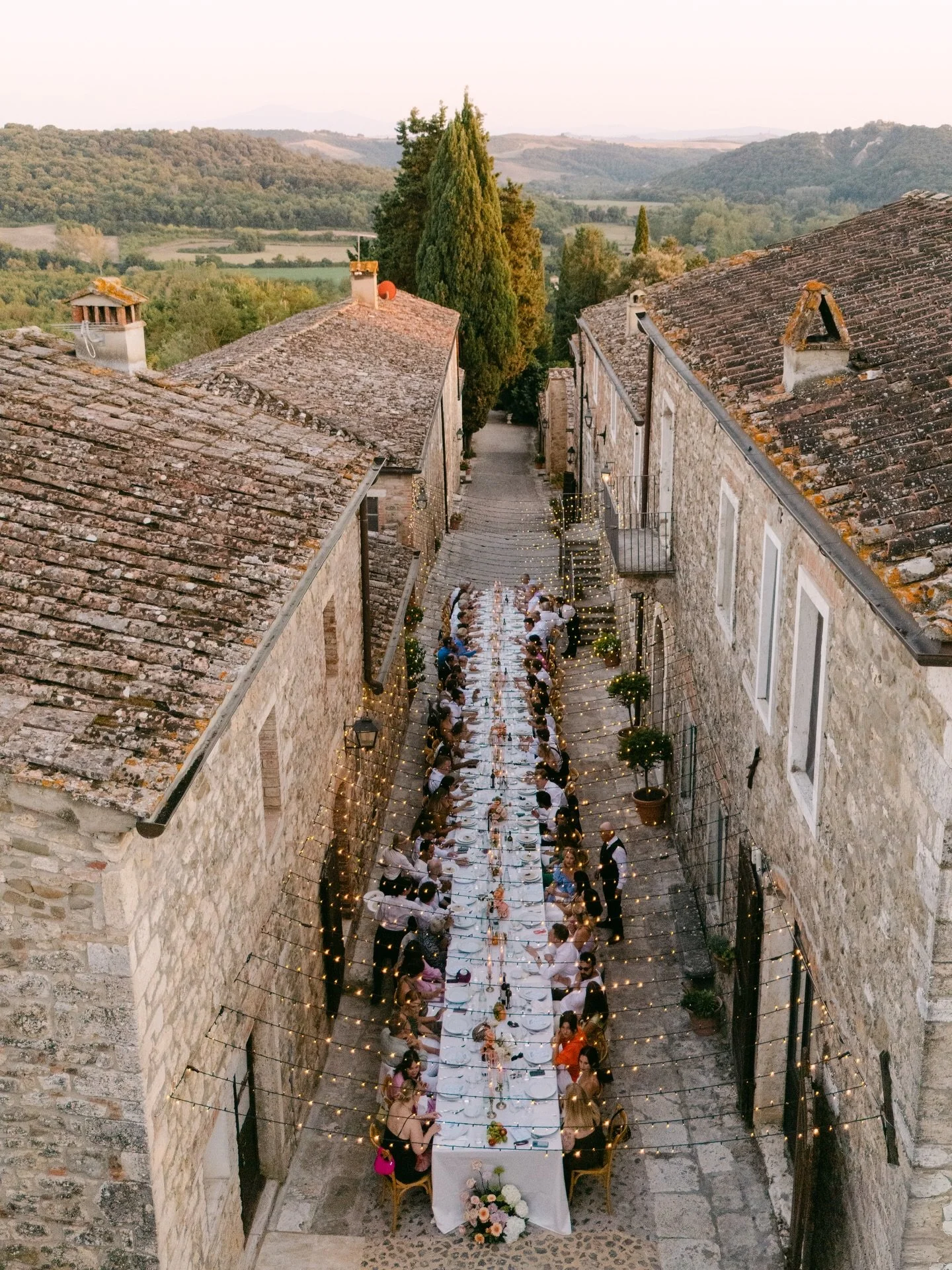 Long wedding dinner table set between historic stone buildings beneath a fairy light canopy at Borgo Laticastelli, Tuscany.