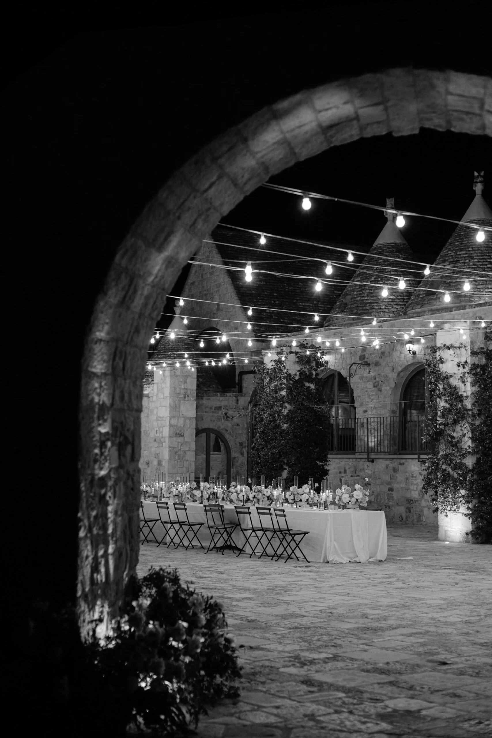 Black and white image of a fairy-lit dinner with stone archway at a historic masseria wedding venue in Puglia, Italy.