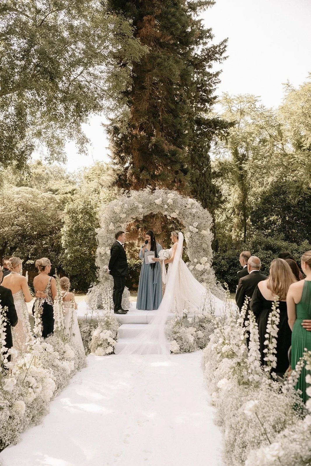 Wedding ceremony along a white floral aisle surrounded by dense green parkland at a centuries-old villa in Palermo, Sicily.
