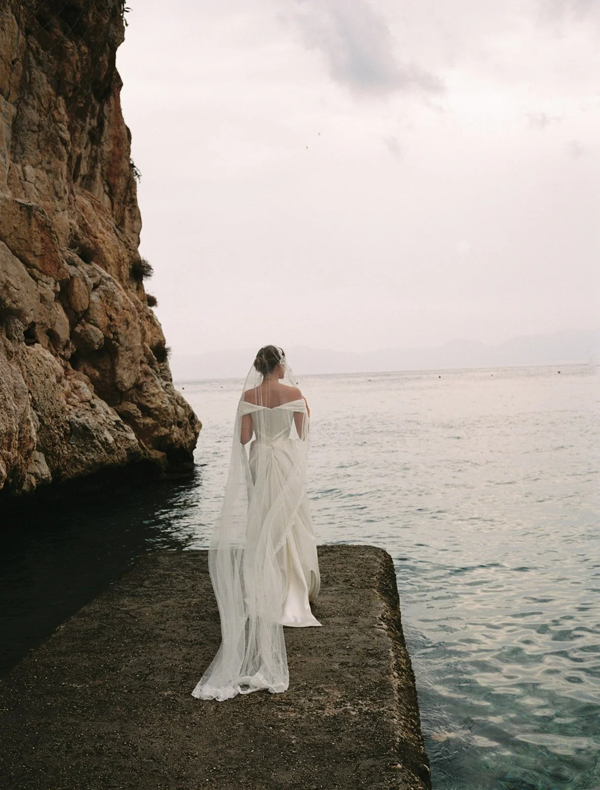 Bride standing on a rocky jetty in Sicily, with the Mediterranean Sea stretching out in front of her.
