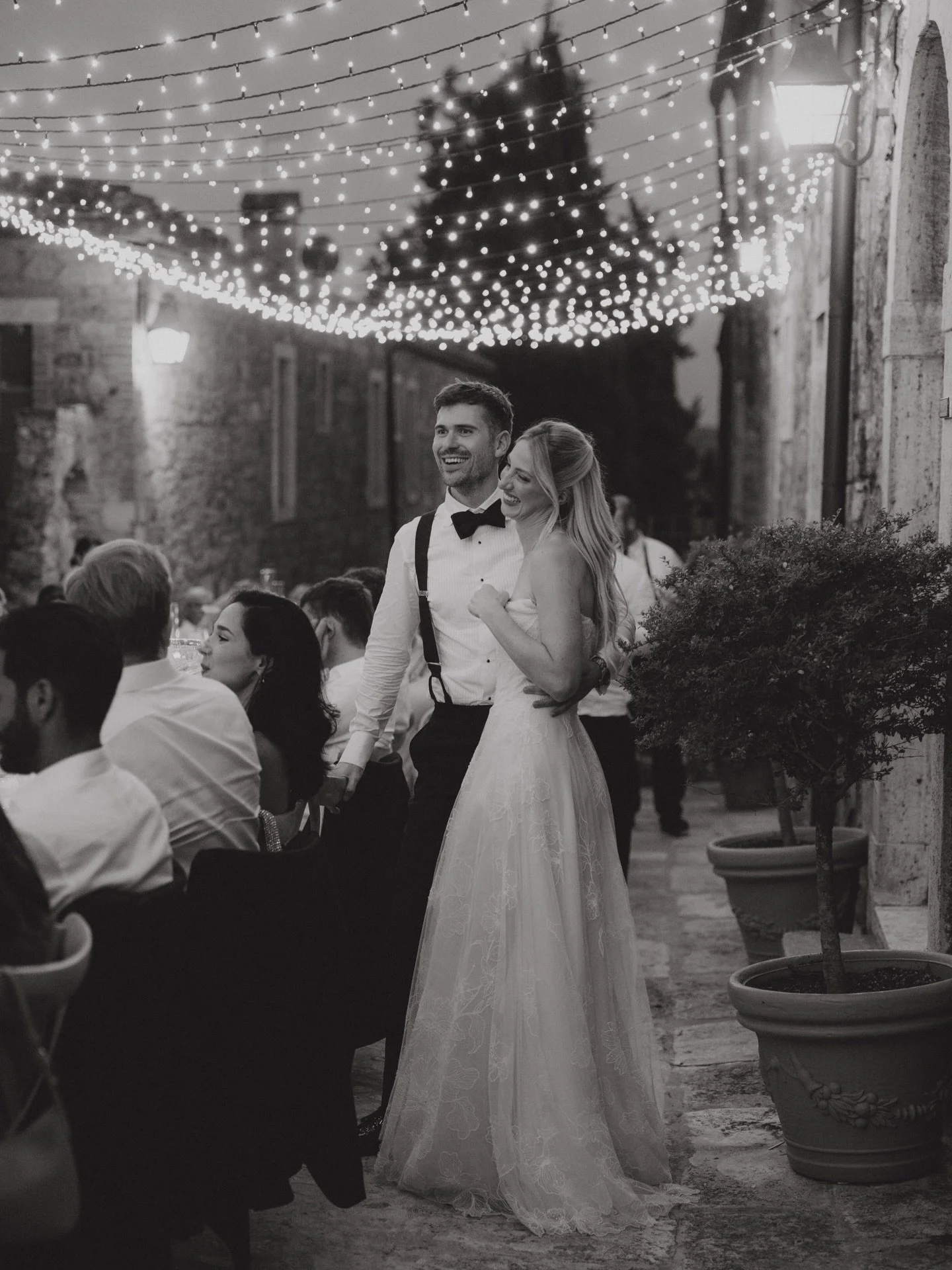 Couple at a wedding dinner beneath a fairy light canopy along an ancient village street in the Crete Senesi, Tuscany.