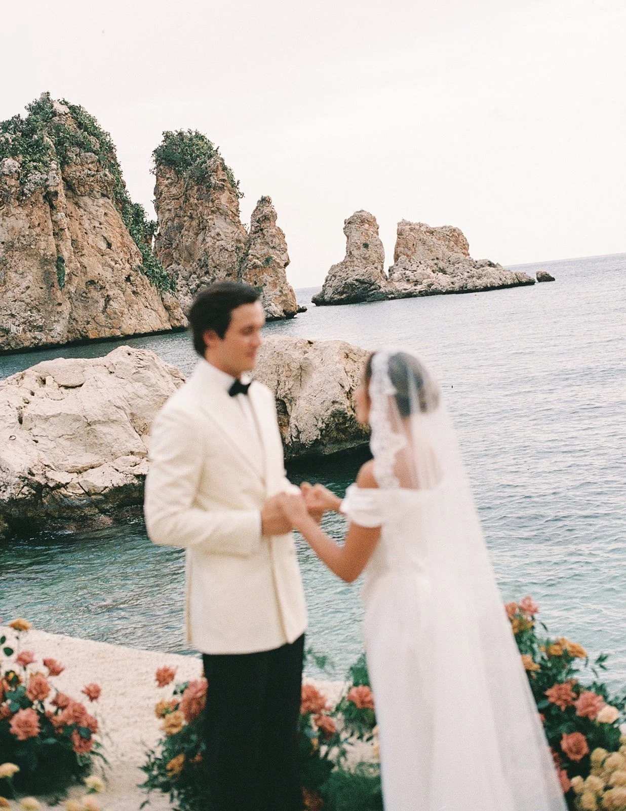 Outdoor wedding ceremony by the sea in Sicily, with a floral arch and dramatic faraglioni sea stacks in the background.