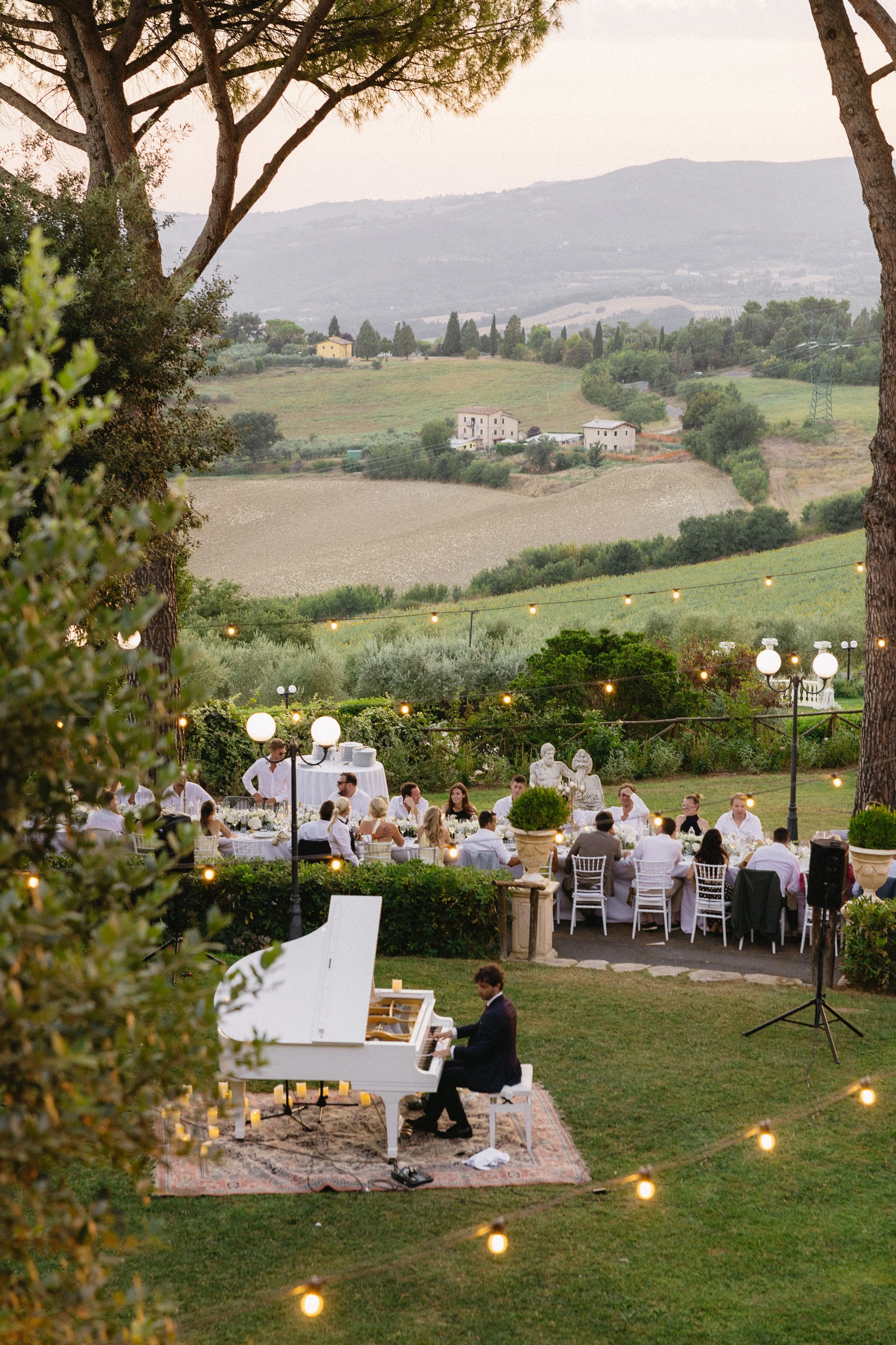 Outdoor wedding dinner at dusk at a historic Umbrian monastery venue, with a pianist performing beside long table.