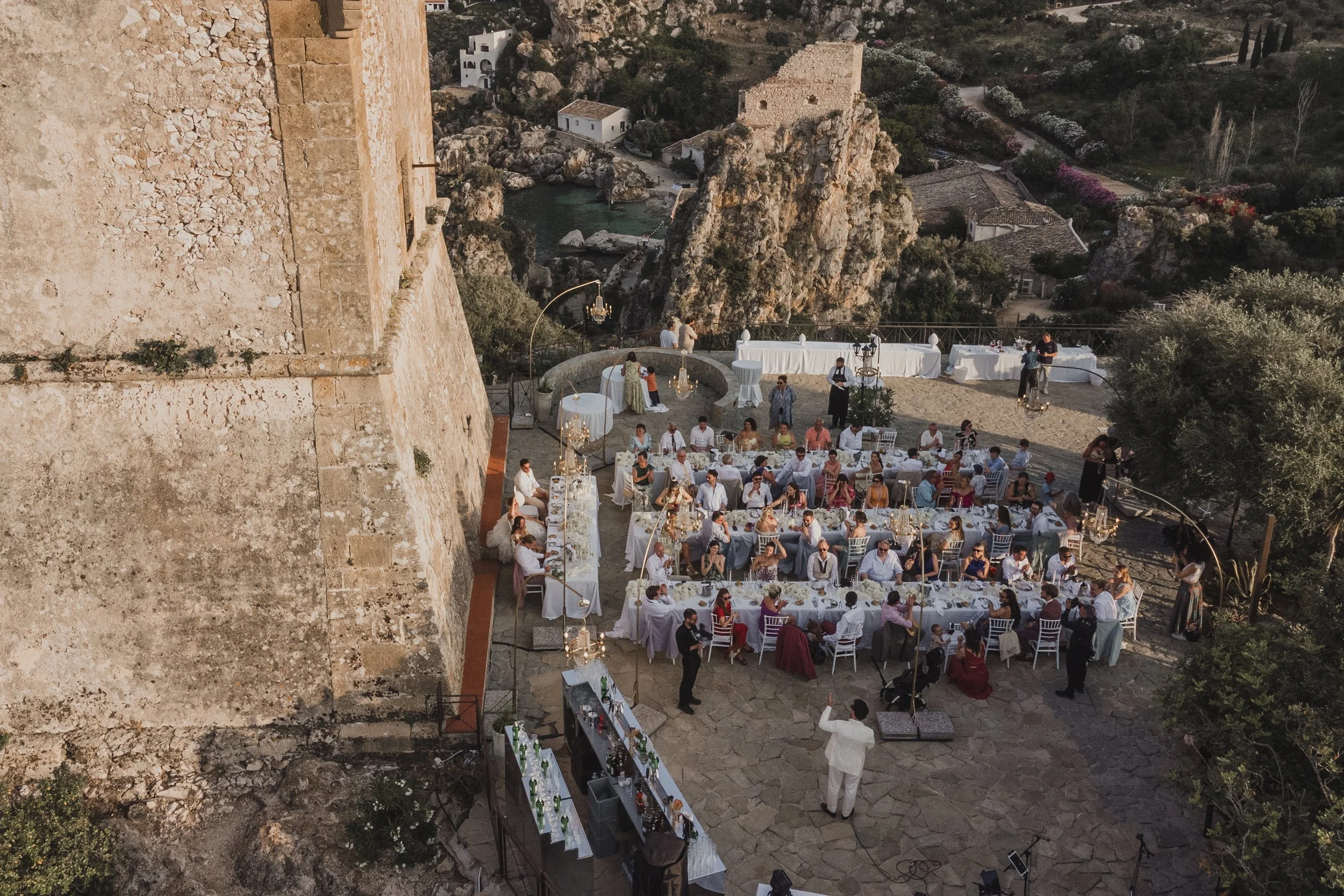 View of a wedding dinner set along long tables beside a historic clifftop tower in Sicily, with the Mediterranean Sea stretching out below.