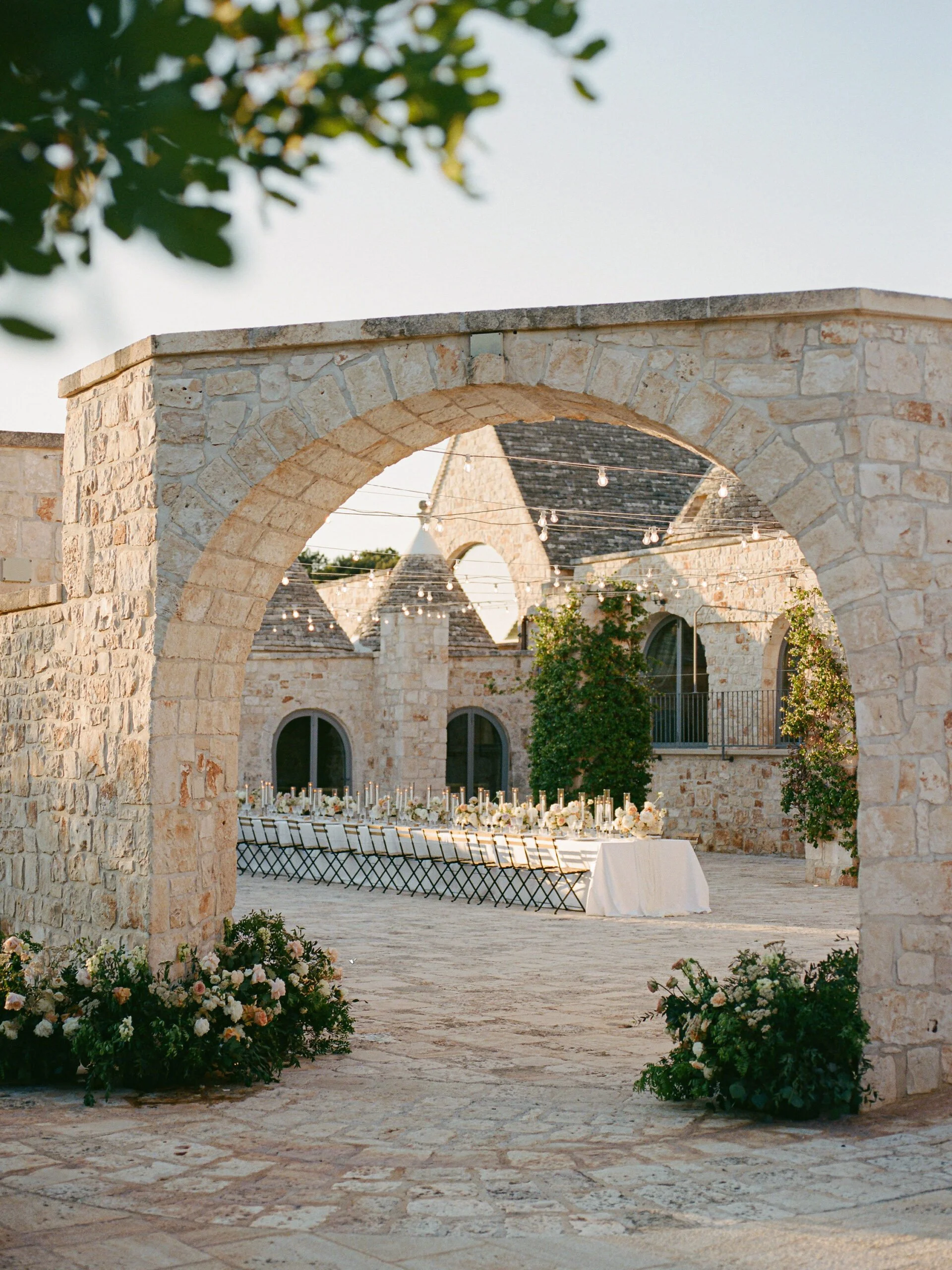 Wedding dinner table at golden hour through a limestone archway at Masseria Grieco, a trulli estate in the Valle d'Itria, Puglia.
