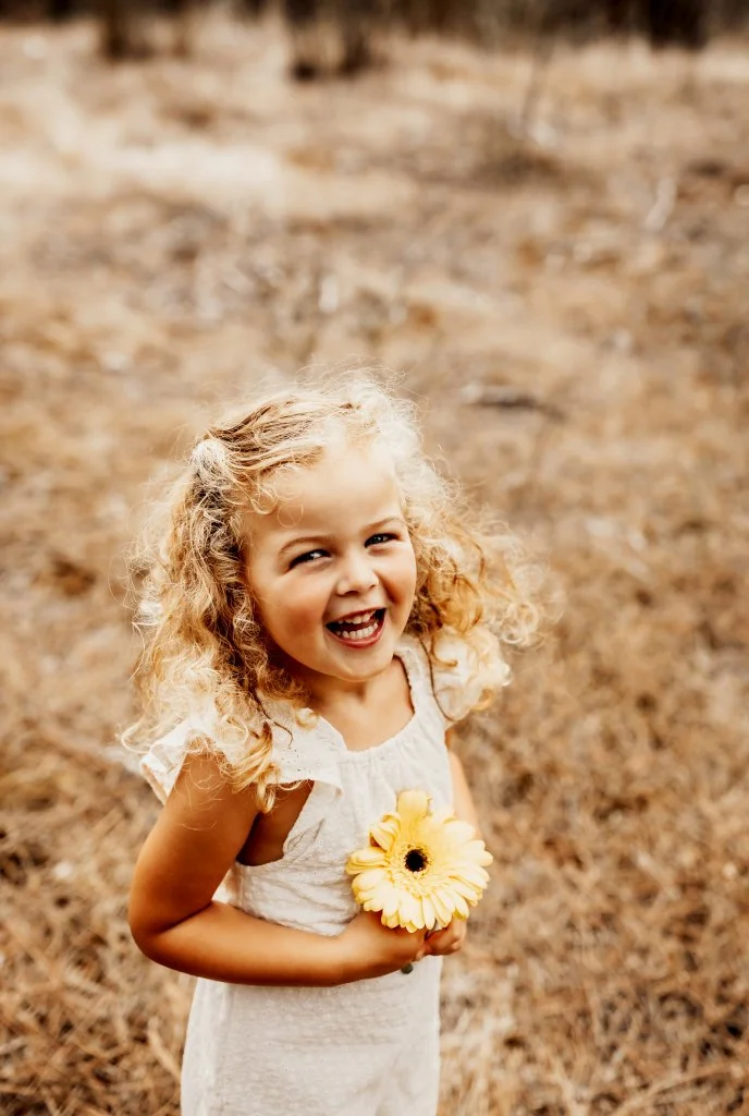 Menina sorridente com cabelo cacheado, segurando uma flor amarela, em um ambiente ao ar livre.