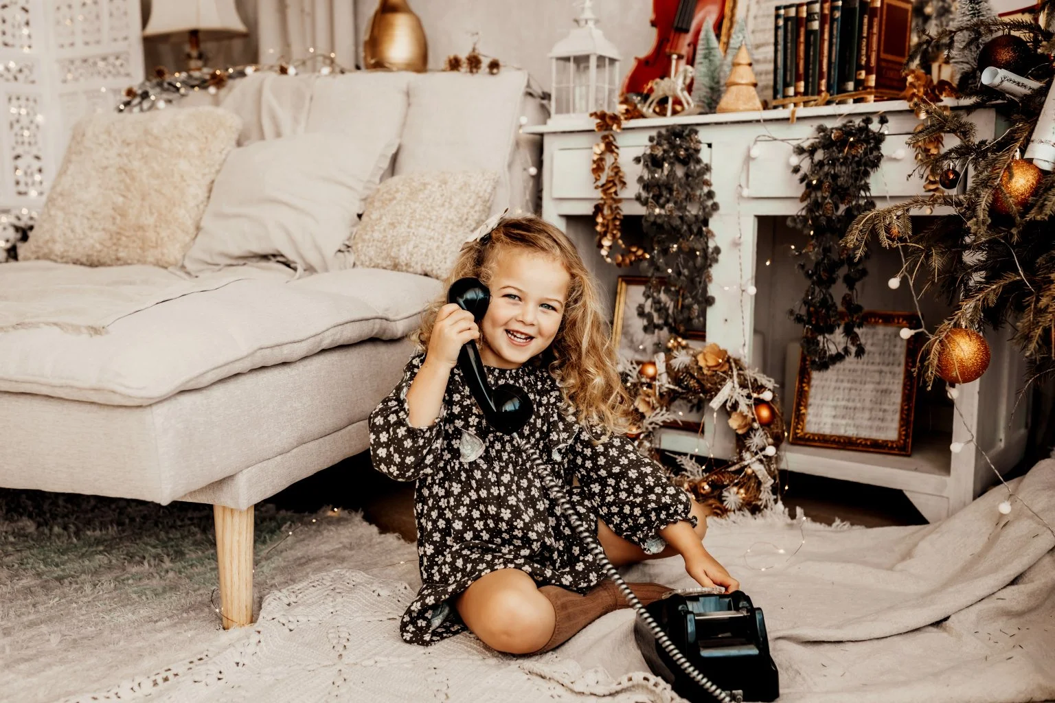 Menina sorridente sentada no tapete, segurando um telefone antigo de disco e brincando com um telefone de brinquedo, em uma sala decorada com enfeites de Natal e uma árvore de Natal decorada.