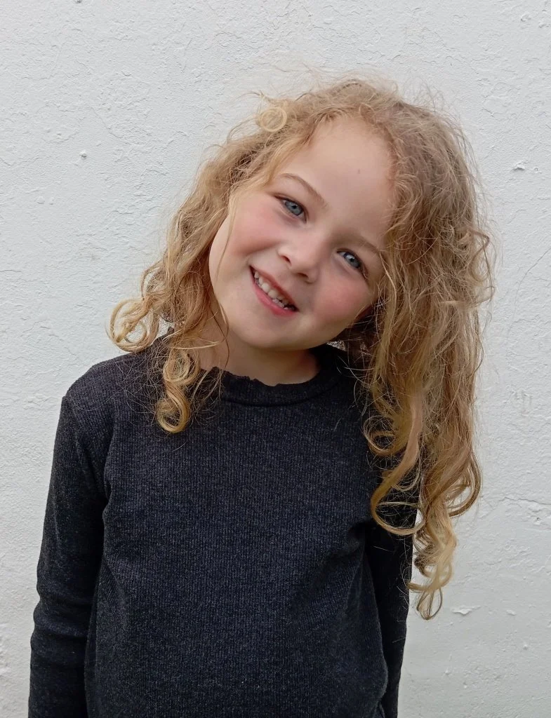 Menina de cabelo cacheado e loiro claro, sorrindo, usando blusa preta, posando ao lado de uma parede branca.
