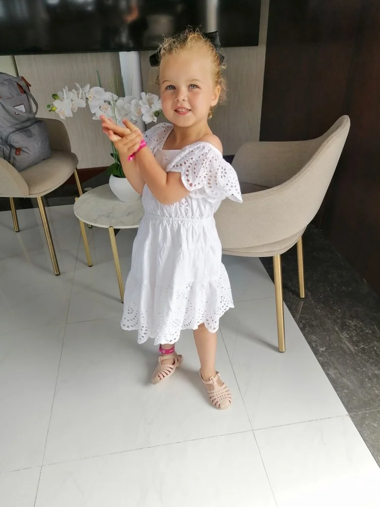 Menina sorridente vestindo vestido branco com detalhes de bordado, usando sandálias rosas, segurando uma flor, em ambiente com cadeiras e uma mesa com uma planta de orquídeas brancas ao fundo.