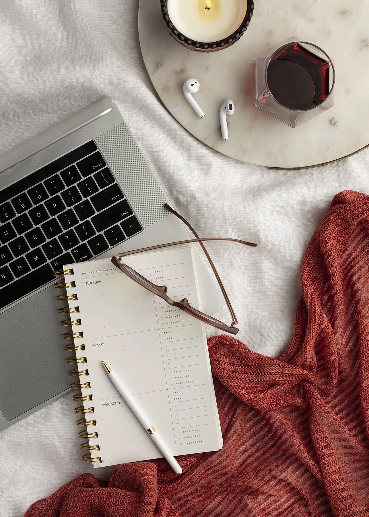 flat lay of a laptop, planner, glasses, pen, earpods, glass of wine, and a red blanket