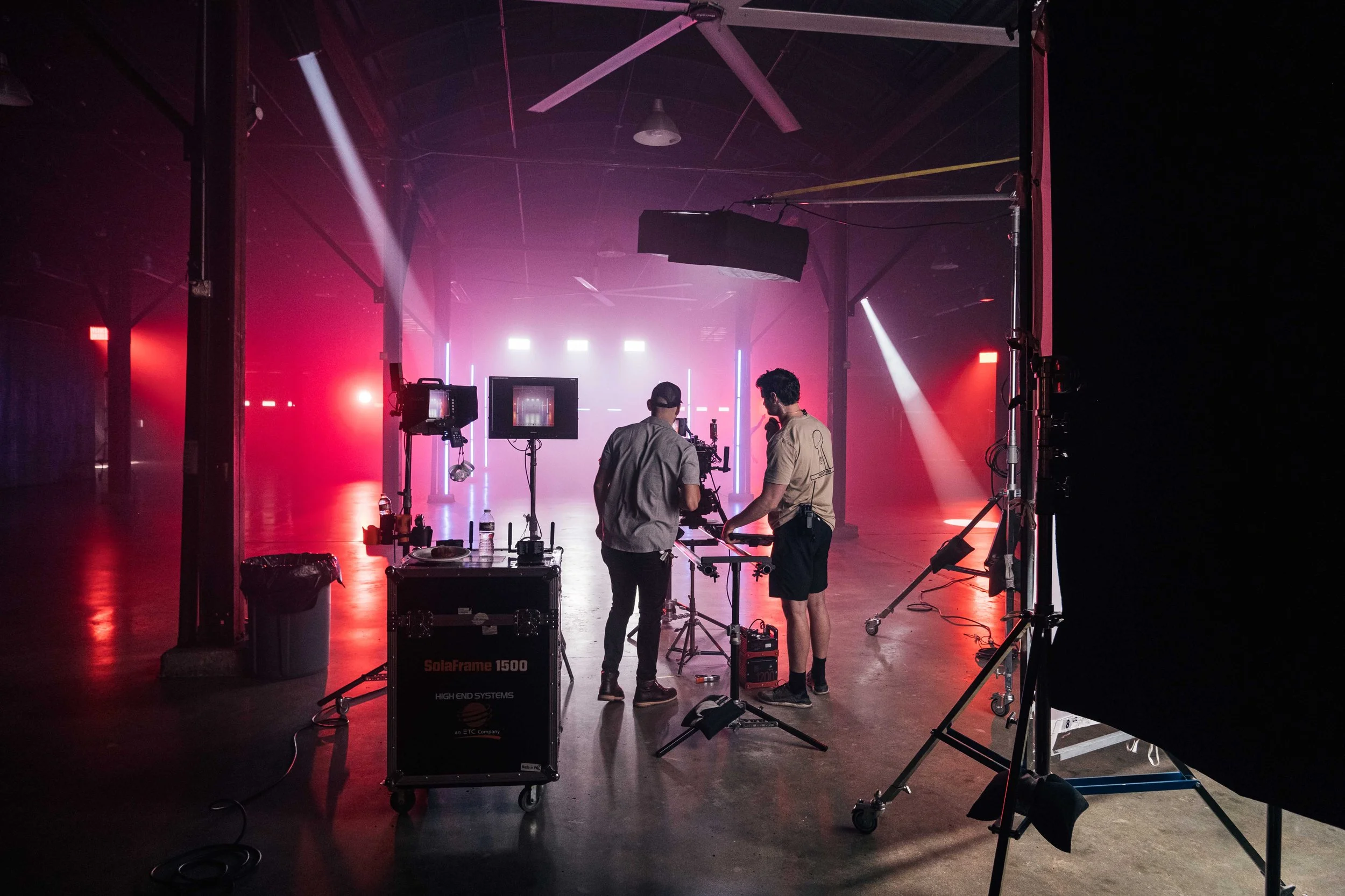 Two crew members working with camera equipment in a dimly lit studio with pink and purple stage lighting and fog.