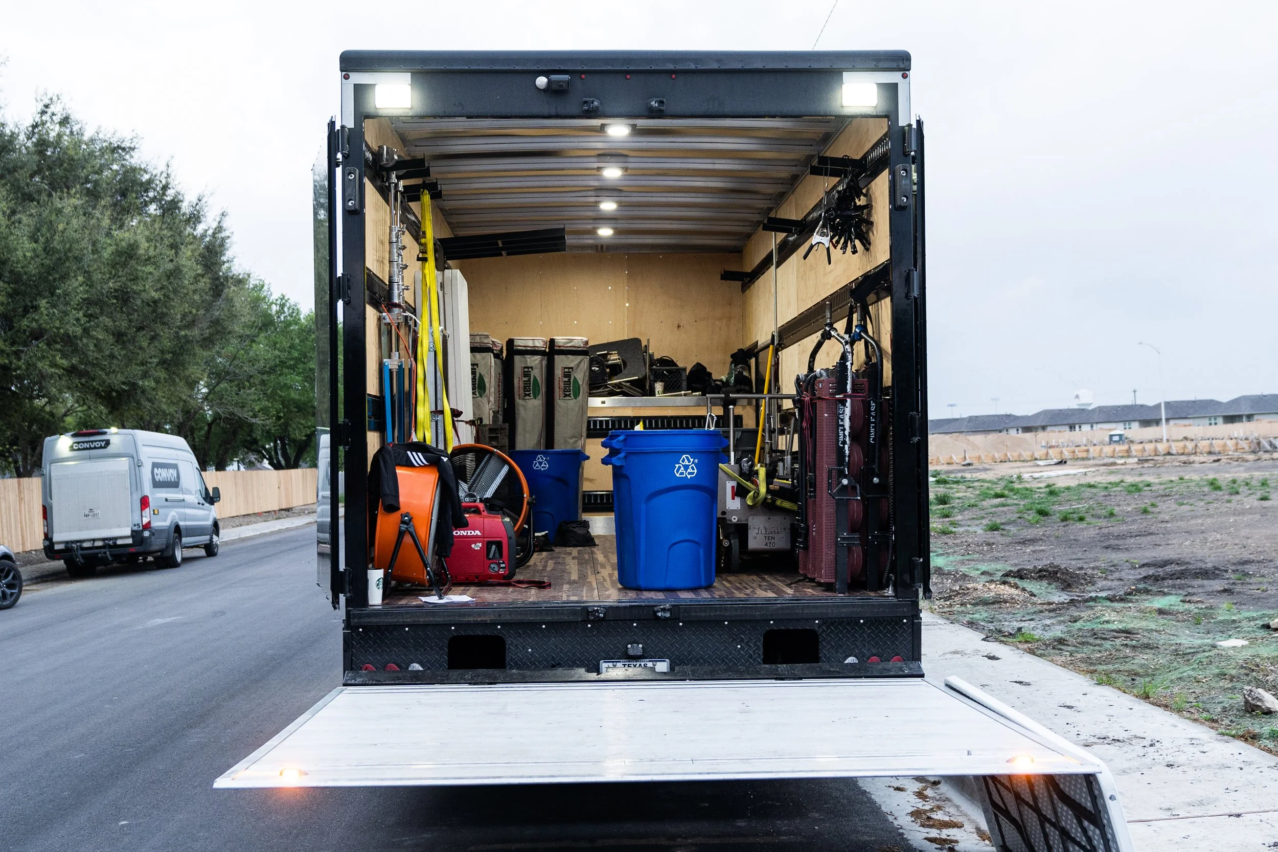 The back view of an open 5-ton truck on a suburban street, showing interior shelves with supplies, tools, and equipment, with a couple of large trash bins in the foreground.