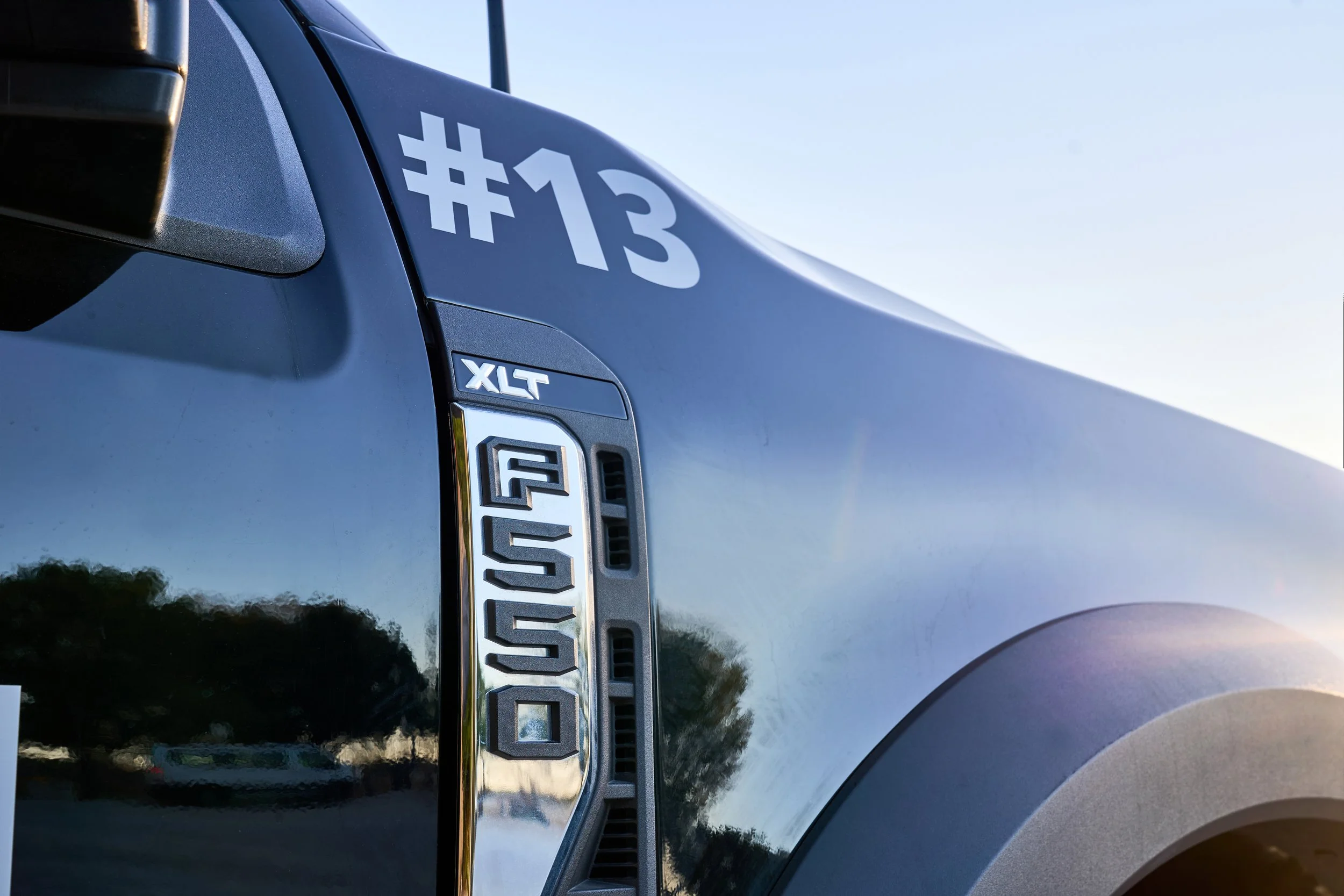 Utility vehicle equipped with various tools and equipment, driving on a dirt path through a wooded area with leafless trees.