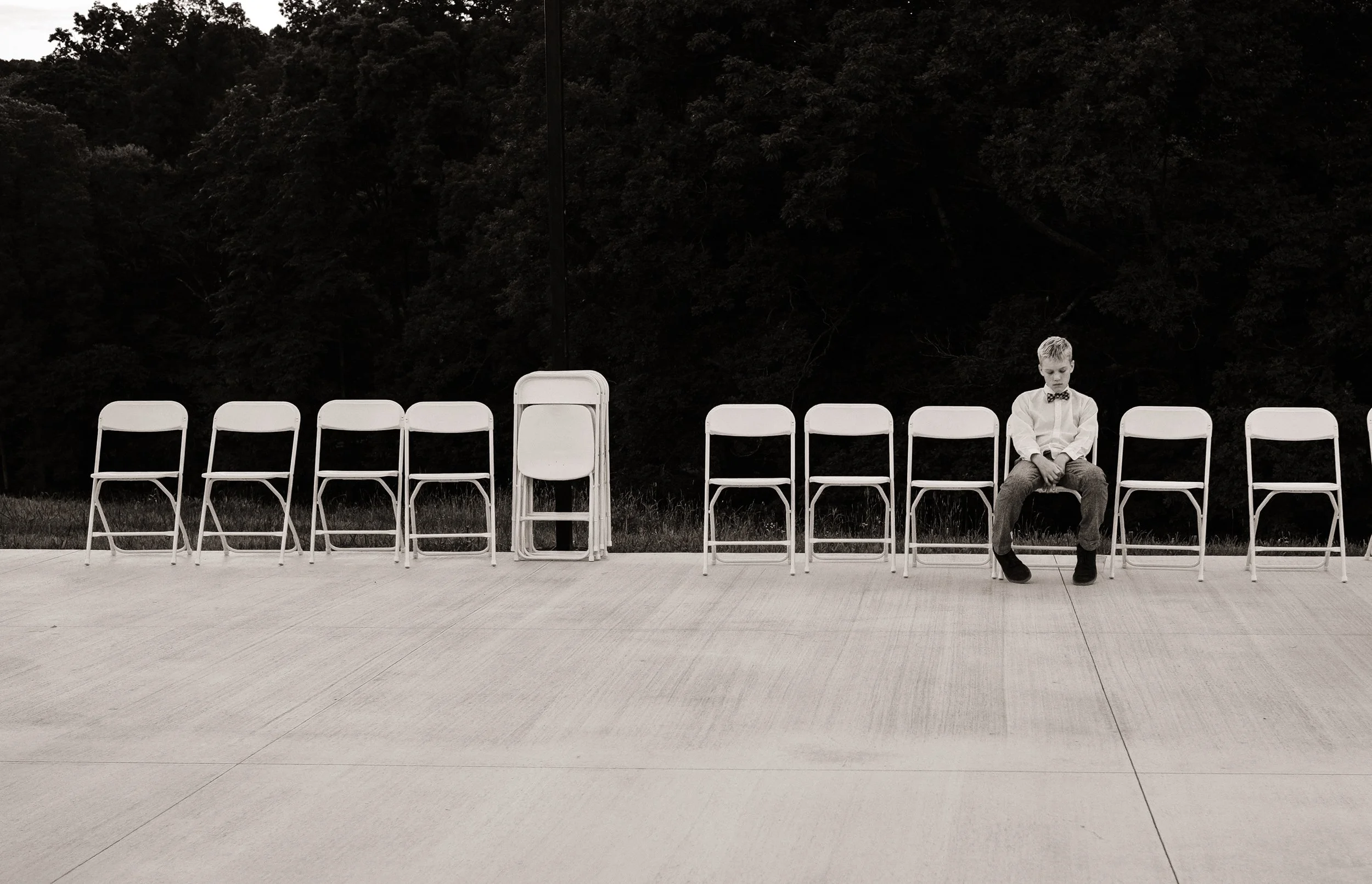 Black and white photo of a young man at a wedding, sitting by himself looking bored.