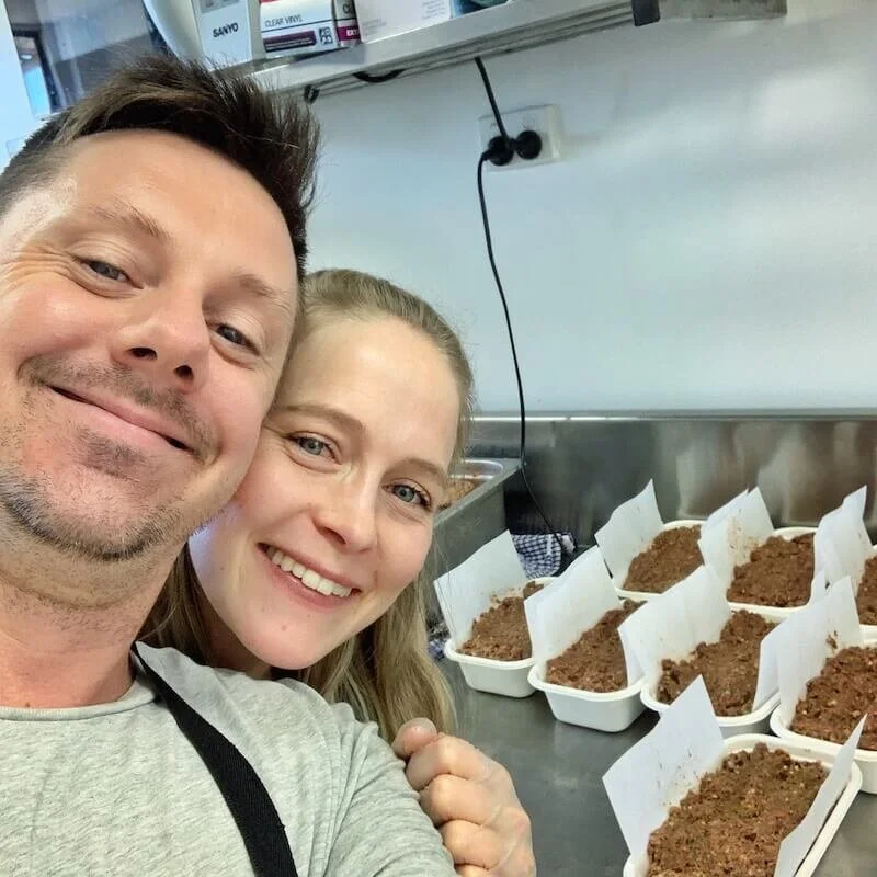 Two people smiling in a kitchen next to several containers of what appears to be soil or planting medium, each with a small paper label, possibly preparing for planting or gardening.