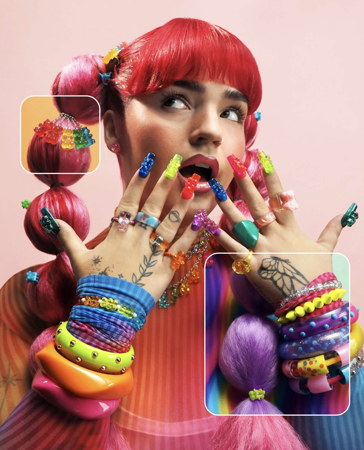 A young woman with bright red hair styled with pink and purple hair ties, wearing colorful rings, bracelets, and nail polish, posing with her hands near her face and mouth open in surprise or excitement, against a pink background.