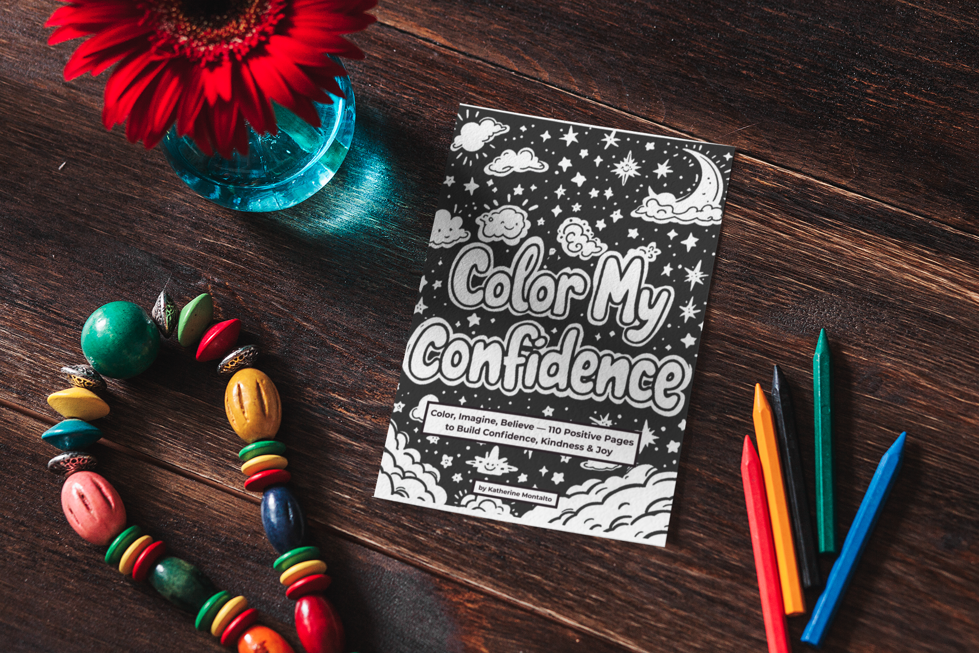 Book titled 'Color My Confidence' on a wooden table, surrounded by colored markers, a beaded necklace, and a vase with a red flower.