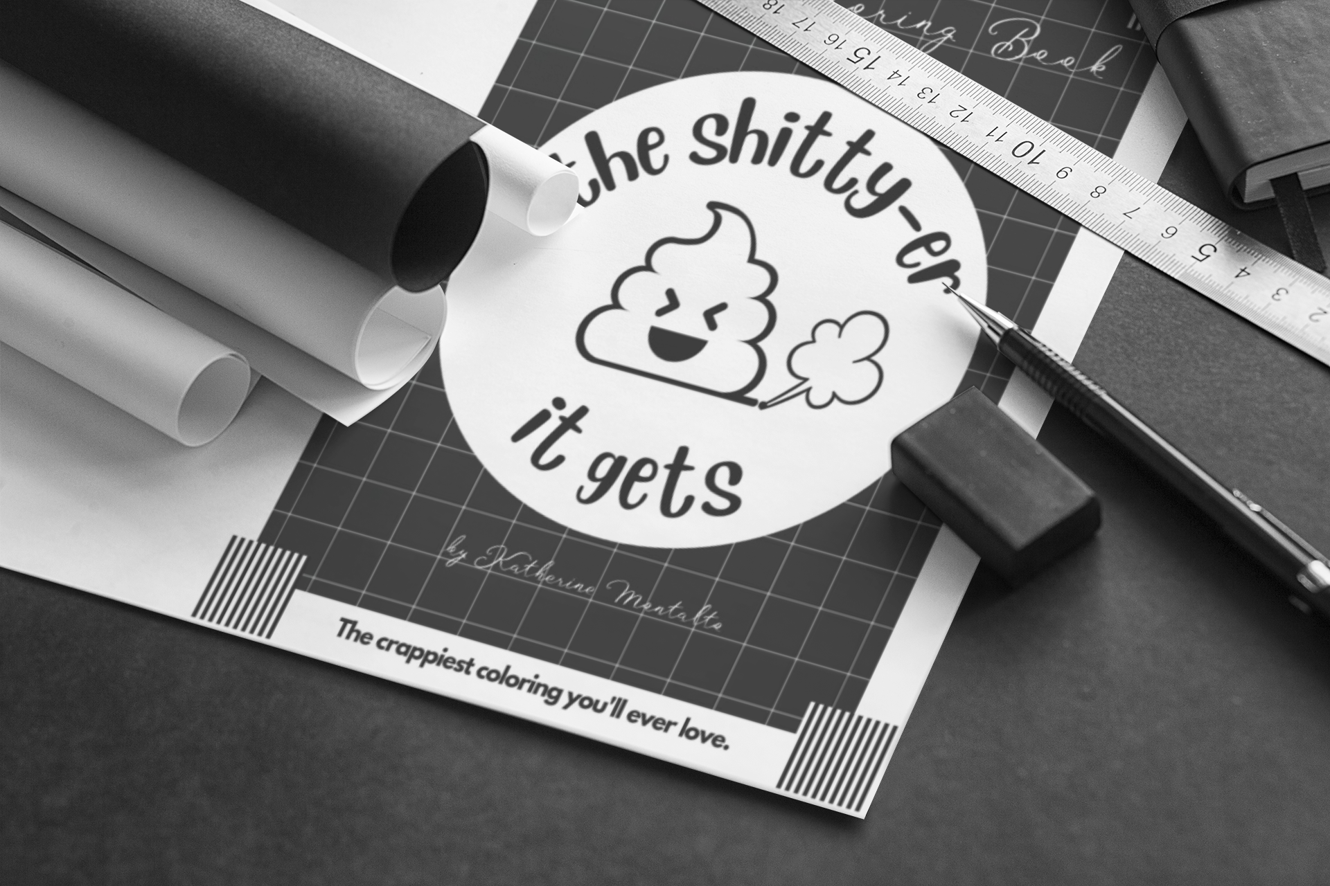 Black and white photo of a circular sticker with a cartoon poop emoji and text that reads "the shitty-er it gets." The sticker is on a workspace with various papers, a ruler, a pen, and an eraser around it.