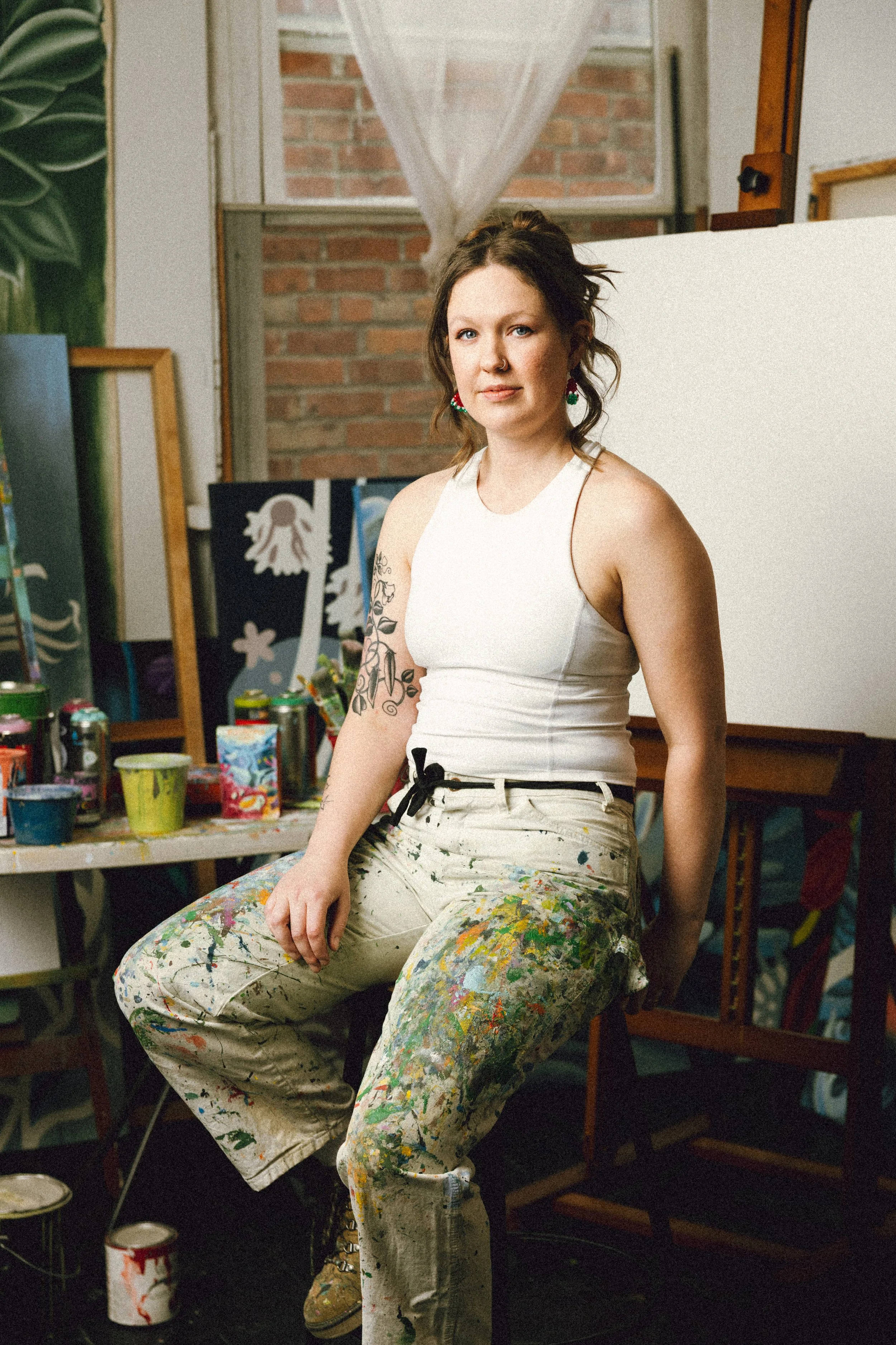 A young woman with tattoos wearing paint-splattered overalls, sitting in an art studio surrounded by paint cans and artwork.