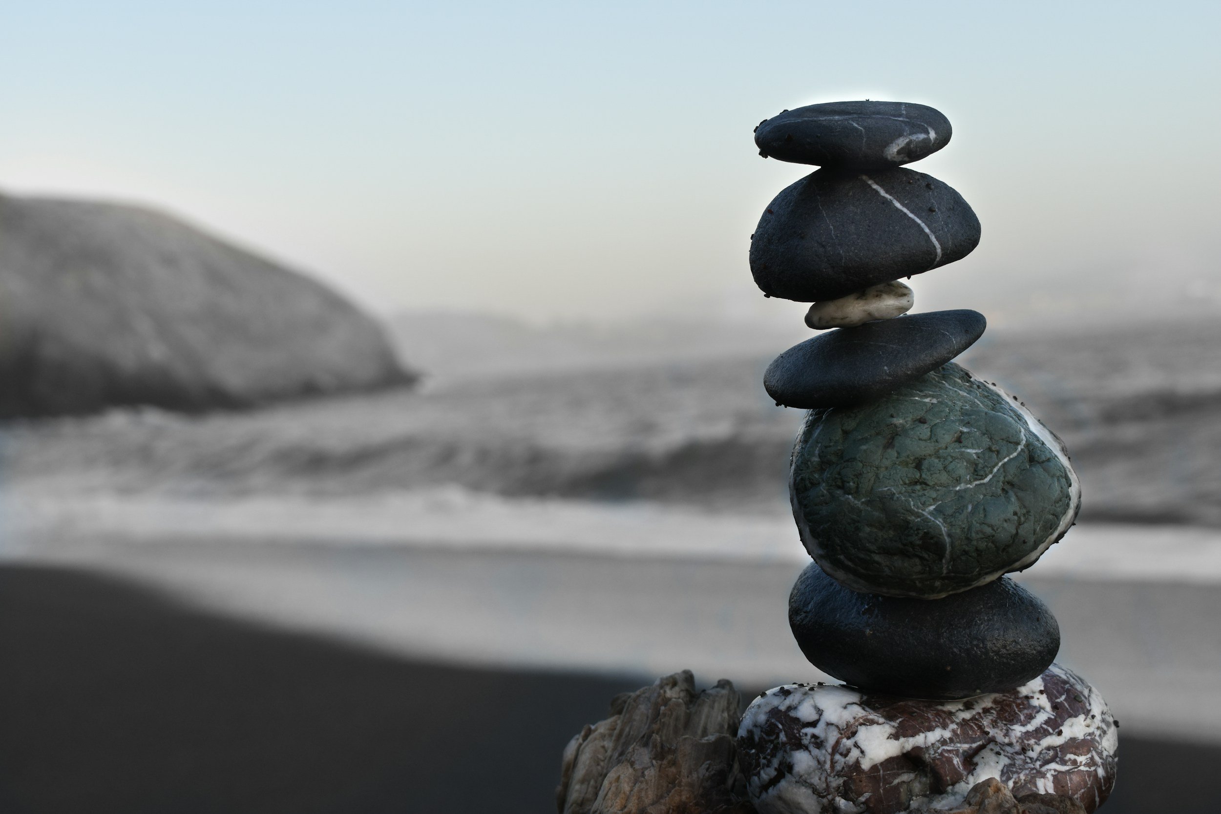 A stack of six smooth stones balanced on a larger rock by the ocean, with waves and a rocky island in the background — symbolizing balance, stability, and the steady foundation created through trauma-informed care and mindful organizational practices