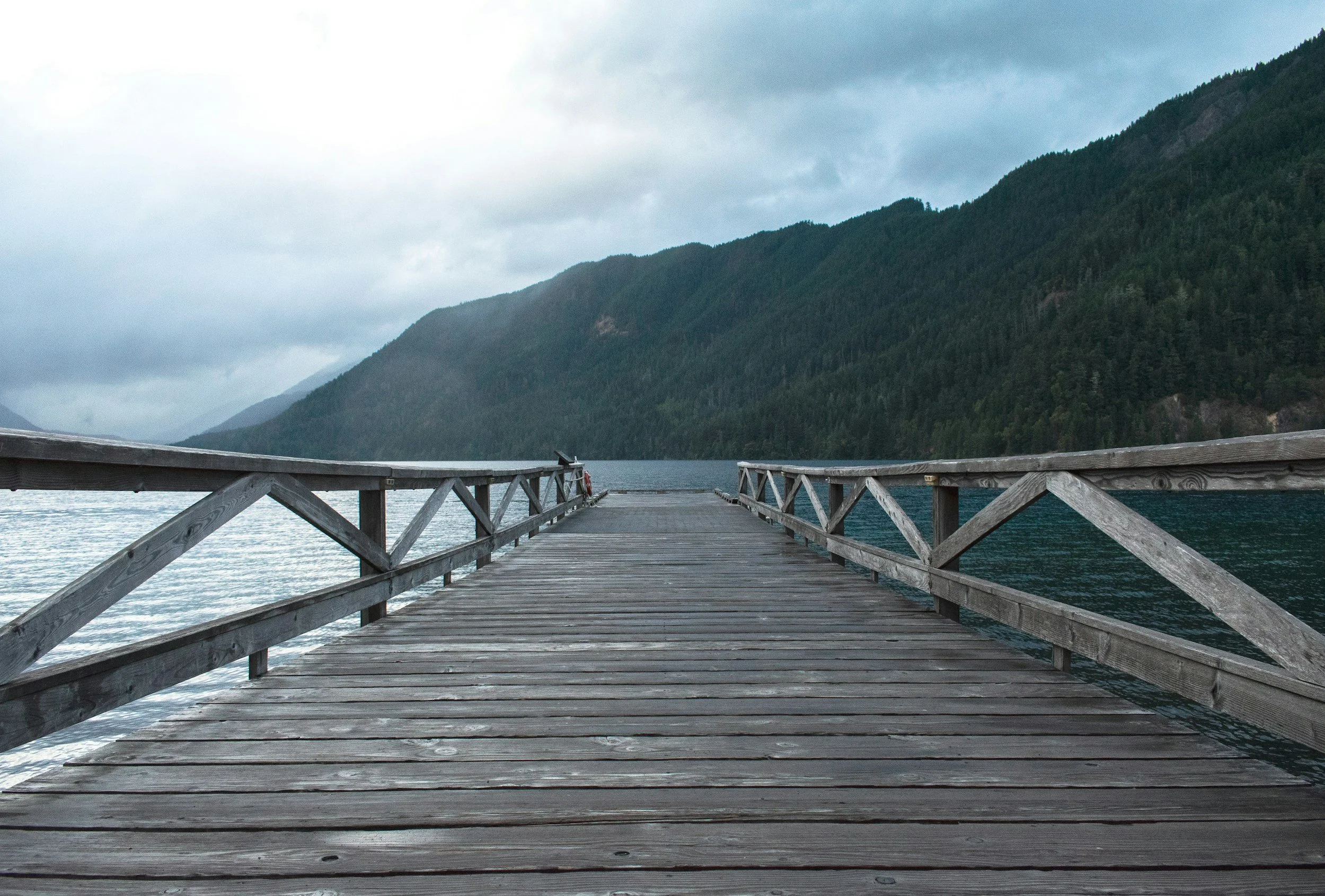 A wooden dock extending over calm water toward tree-covered mountains under a cloudy sky — representing the bridge-building work of Cultural Intelligence, connecting people and perspectives across differences to foster understanding and inclusion.