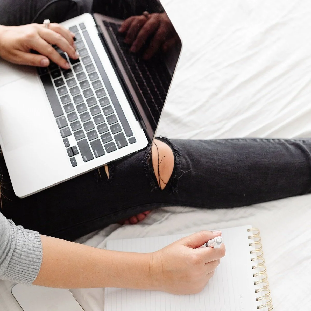 Person sitting on a white surface with a laptop on their lap, holding a notebook and pen, wearing ripped black jeans and a gray sweater.
