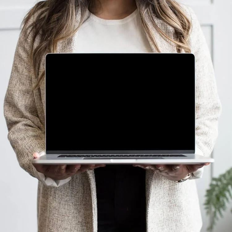Person holding a laptop with a black screen in front of them, dressed in a beige coat and white shirt.