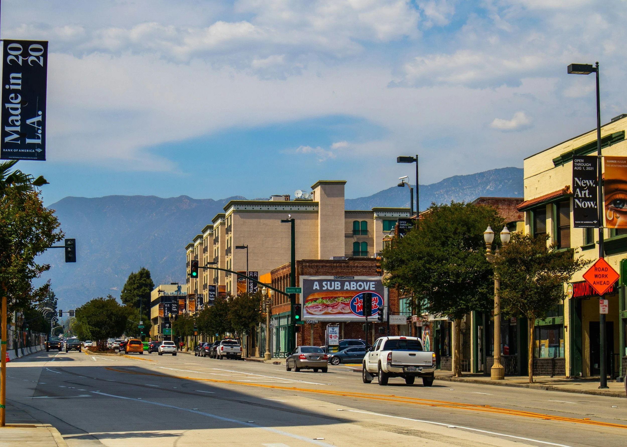 Urban street scene with cars, buildings, and a billboard promoting sandwiches. Banners along the street say "Made in L.A. 2020." Mountains visible in the background under a cloudy sky.